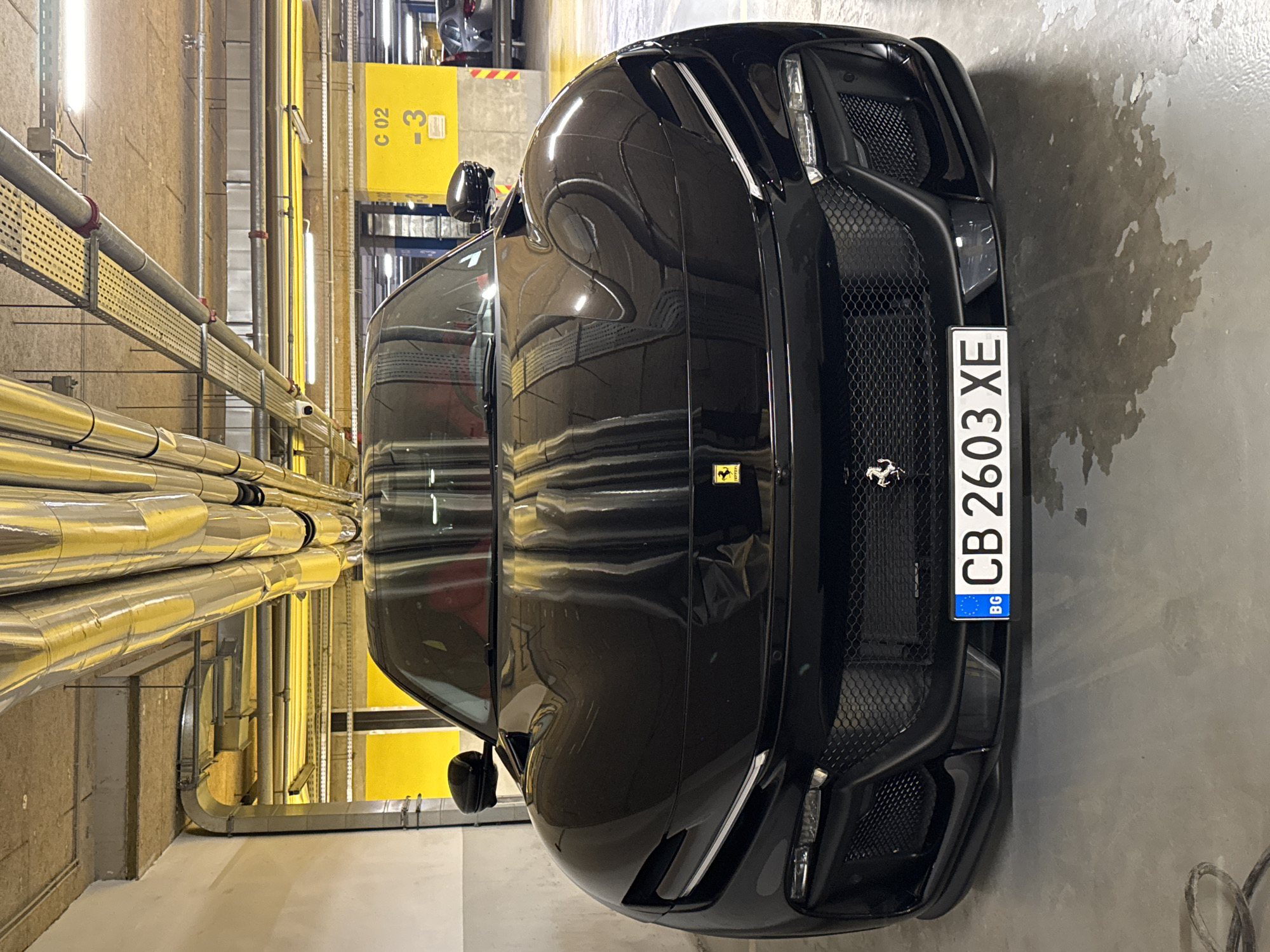 A black Ferrari is parked in a brightly lit indoor parking garage in Sofia, Bulgaria. The car is positioned perpendicular to the camera. A yellow sign with the text "C 02 -3" is visible in the background. The license plate number on the car is CB 2603 XE. Large metal pipes run along the ceiling of the garage. The parking garage is located in Sofia, Bulgaria as noted by the BG on the numberplate.