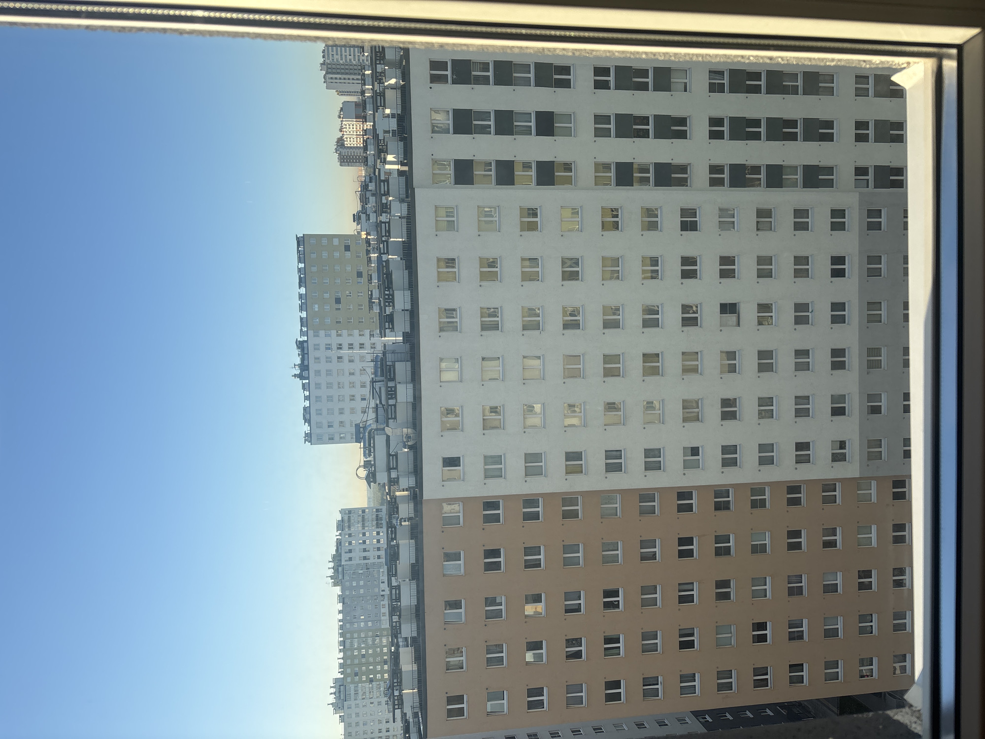 The image shows a view of multiple high-rise apartment buildings from within another building, likely through a window. The sky is visible to the left of the buildings and is blue. The buildings are primarily white and brown with repeating window patterns. Some buildings have balconies. The view appears to be from a high vantage point within the city. The exact location is in an unknown location in Russia.