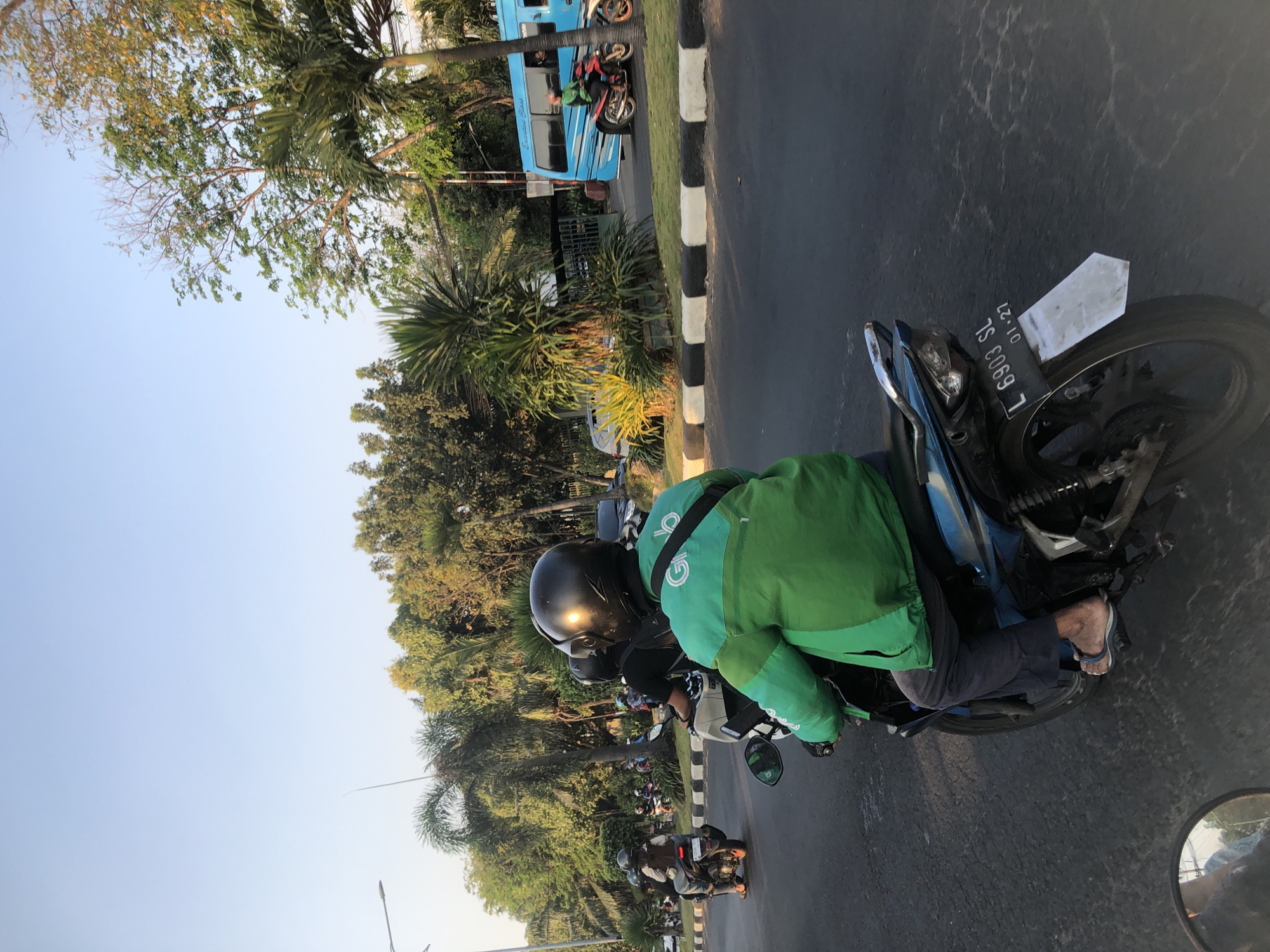 A GrabFood delivery driver, wearing a green jacket and black helmet, rides a motorcycle on a paved road. The driver's jacket has the Grab logo on the back. The motorcycle has a license plate that reads "L 6903 SL 01-27". In the background, there are trees, a blue bus, and other vehicles. The scene appears to be in Candi, Indonesia, given the typical setting for such a delivery service.