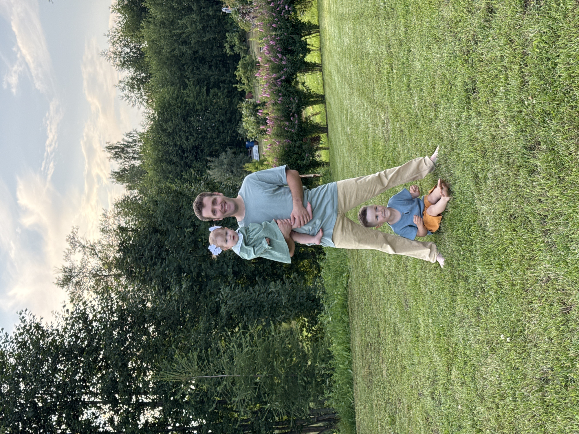 A man stands on a grassy lawn in a rural setting, possibly in the city of Garevaya, Russia. He is holding a baby girl in a green dress and white bows in her hair. A young boy in a blue shirt and orange shorts sits on the grass in front of him. The man is wearing a light blue t-shirt and beige pants. Behind them are trees and flowering bushes. The sky is cloudy.