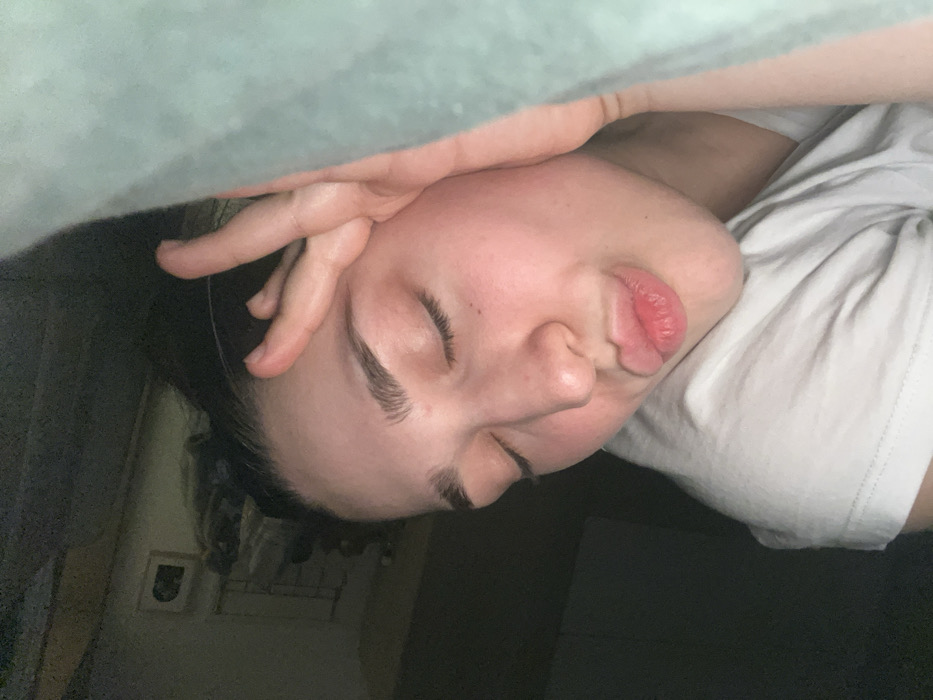 A close-up, angled shot depicts a person's face with eyes closed, a hand resting on their forehead, and pursed lips. They are partially obscured by a teal fabric from above. The individual wears a white t-shirt. The background shows glimpses of a dimly lit room, including a wall and a dark-colored surface. The overall impression is one of rest or sleep.