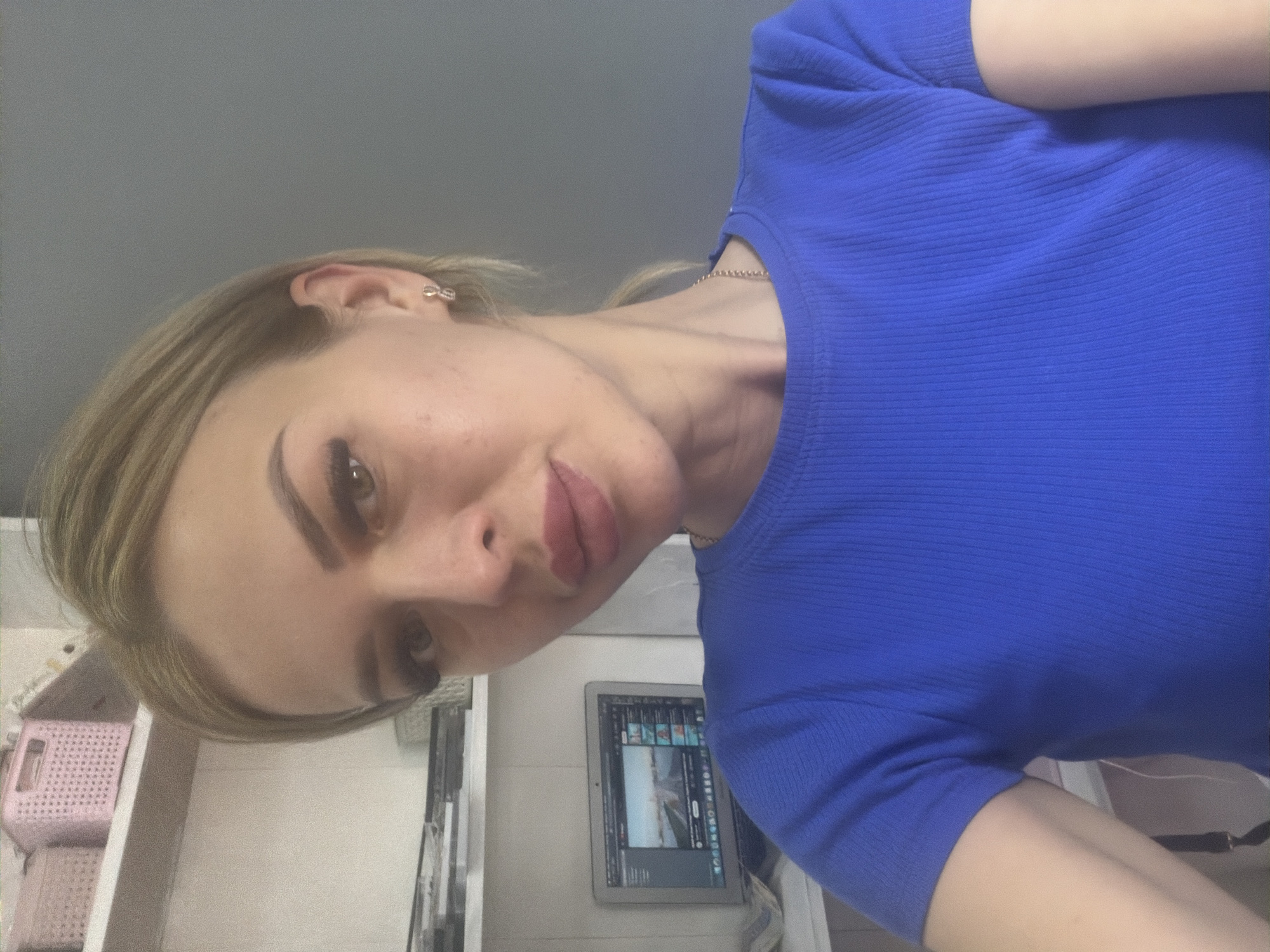 A woman with blonde hair and makeup is taking a selfie in a room. She is wearing a blue t-shirt and a necklace. Behind her, there are shelves with pink baskets and a laptop displaying a video. The room appears to be an indoor setting, possibly an office or living space. No information is available to confirm the location in Kyiv, Ukraine.