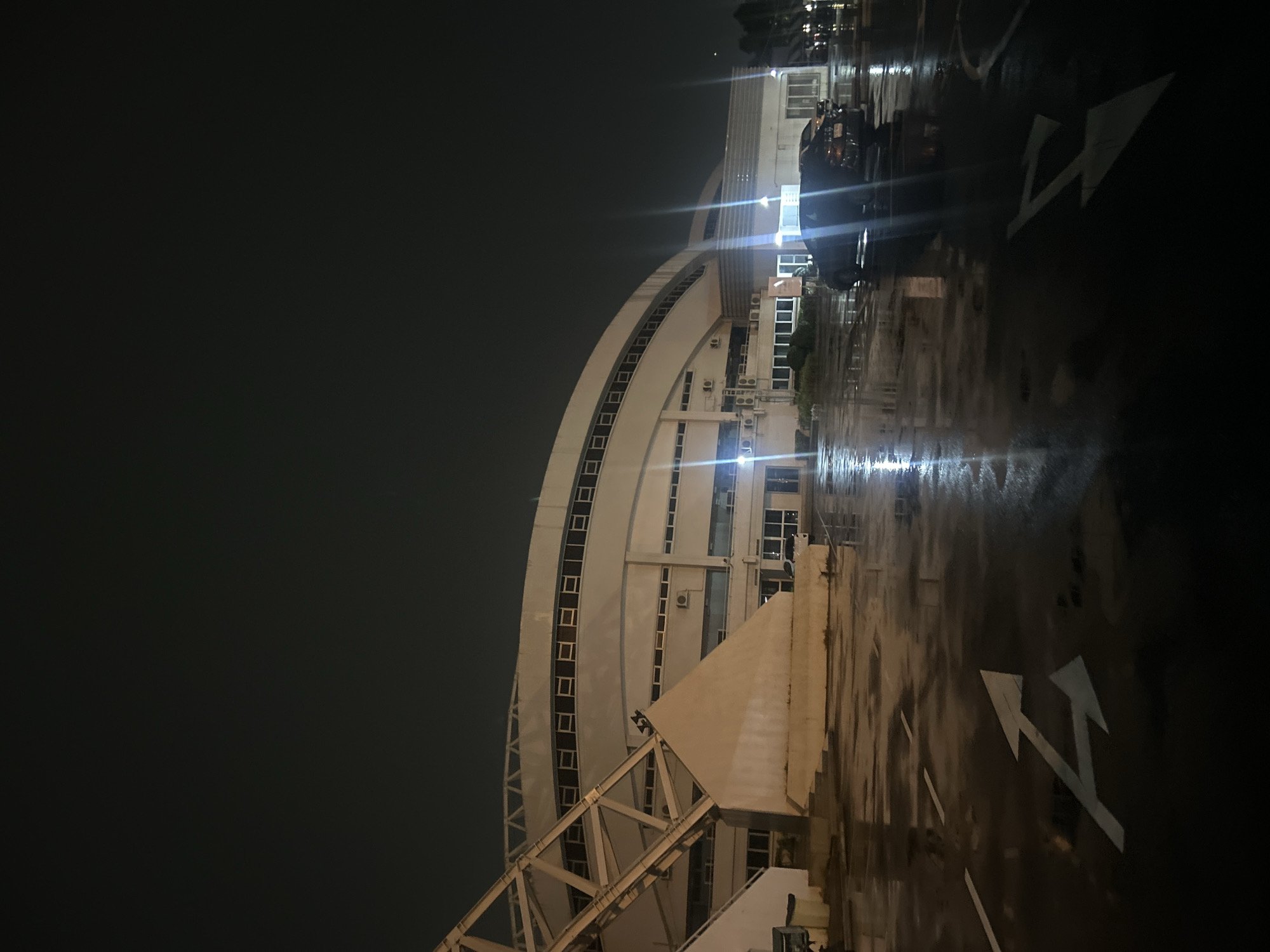 This image depicts a wet, outdoor parking lot at night in front of a large, curved building with a white facade and dark windows. The building appears to be a stadium or arena, with a prominent, angular, white support structure on the left. The parking lot surface is dark and reflective, indicating it has recently rained. White directional arrows are painted on the ground. Several cars are parked in the lot, including a dark-colored sedan in the foreground and another vehicle behind it. Lights from the building and possibly streetlights cast reflections on the wet pavement. The sky is dark and overcast. The scene is located in Tha Khlong Town Municipality, Thailand.