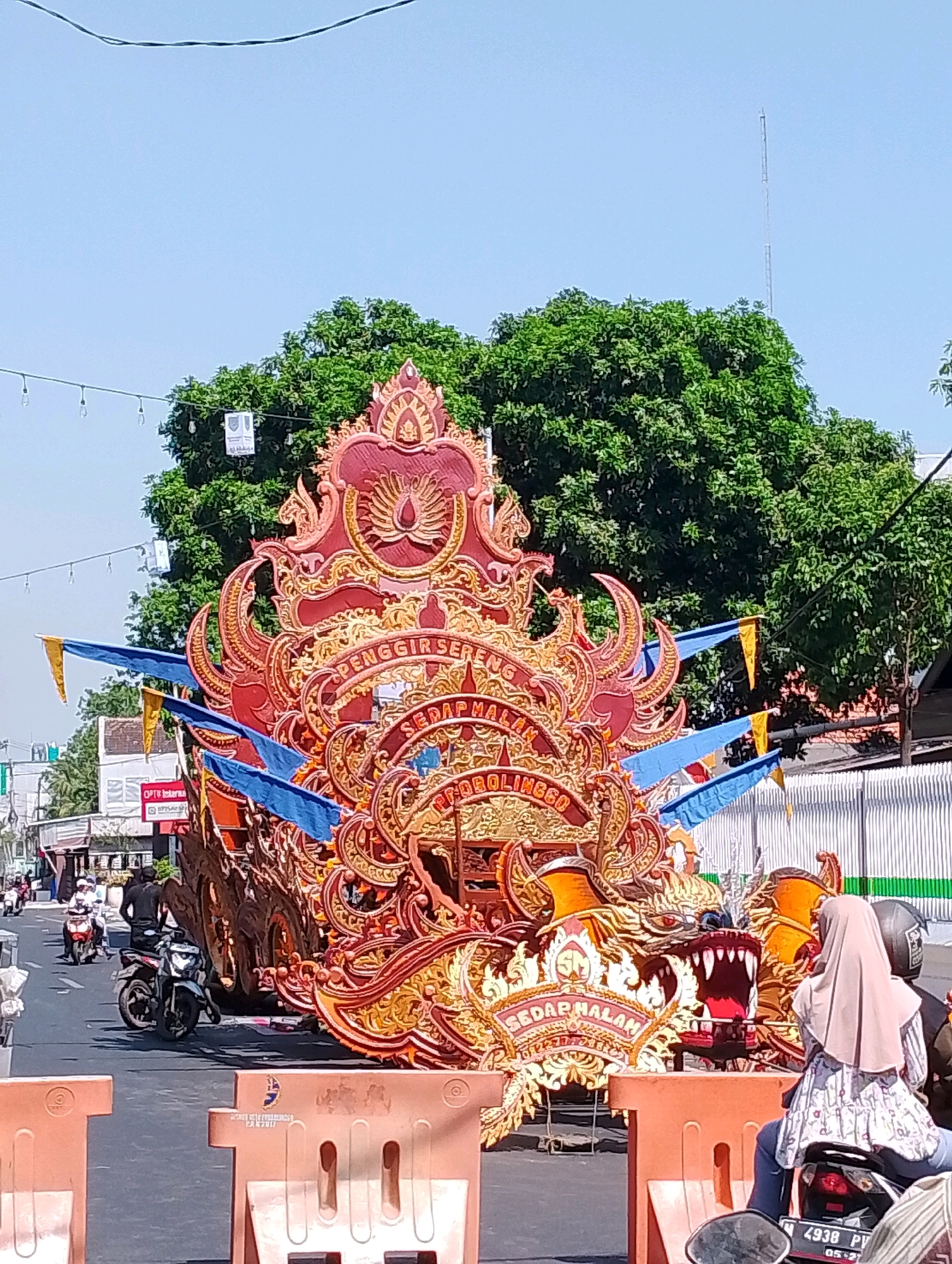The image depicts a vibrant parade float resembling a dragon, adorned with intricate gold and red ornamentation, moving down a city street in Probolinggo, Indonesia. The float is decorated with blue and yellow flags, and inscribed text, including "PENGGIR SEREN" and "SEDAP MALAM PROBOLINGGO". In the foreground, orange traffic barriers line the road, and a person wearing a light-colored hijab is seated on a motorcycle, facing away from the camera. Several other motorcycles and people are visible further down the street, suggesting an ongoing event. The scene is bathed in bright sunlight, with lush green trees lining the background and a clear blue sky overhead. A tall communication tower is visible in the distance. The overall atmosphere is festive and lively.