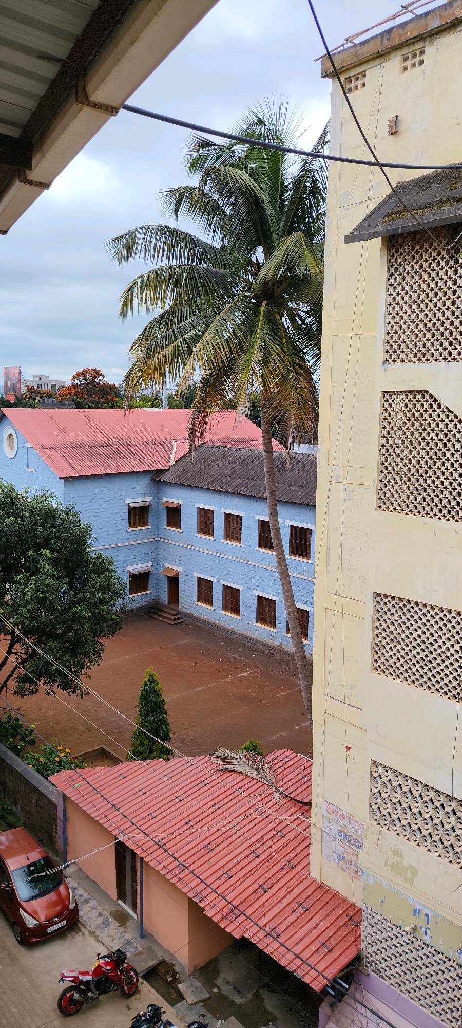 The image depicts a courtyard and buildings, likely a school or administrative complex, under an overcast sky. The scene is set in Belagavi, India, as indicated by the car's license plate. In the foreground, a red car is parked on a paved area next to a single-story building with a red corrugated metal roof. A red motorcycle is parked beside the car.
Behind the single-story building, a two-story building with a light blue facade and a reddish-pink corrugated metal roof is visible. This building has numerous windows with brown frames. A courtyard area in front of it is covered with reddish-brown soil. A tall palm tree grows in front of this blue building, its fronds extending into the frame.
To the right of the blue building, a taller, light yellow building with decorative lattice-work windows dominates the right side of the image. This building appears to be older and shows signs of wear and tear, with some peeling paint.
In the background, beyond the blue building, more structures and trees are visible, including a tree with bright orange flowers. Electrical wires crisscross the scene, strung between the buildings. The overall impression is one of a functional, established complex with elements of both new and older construction.