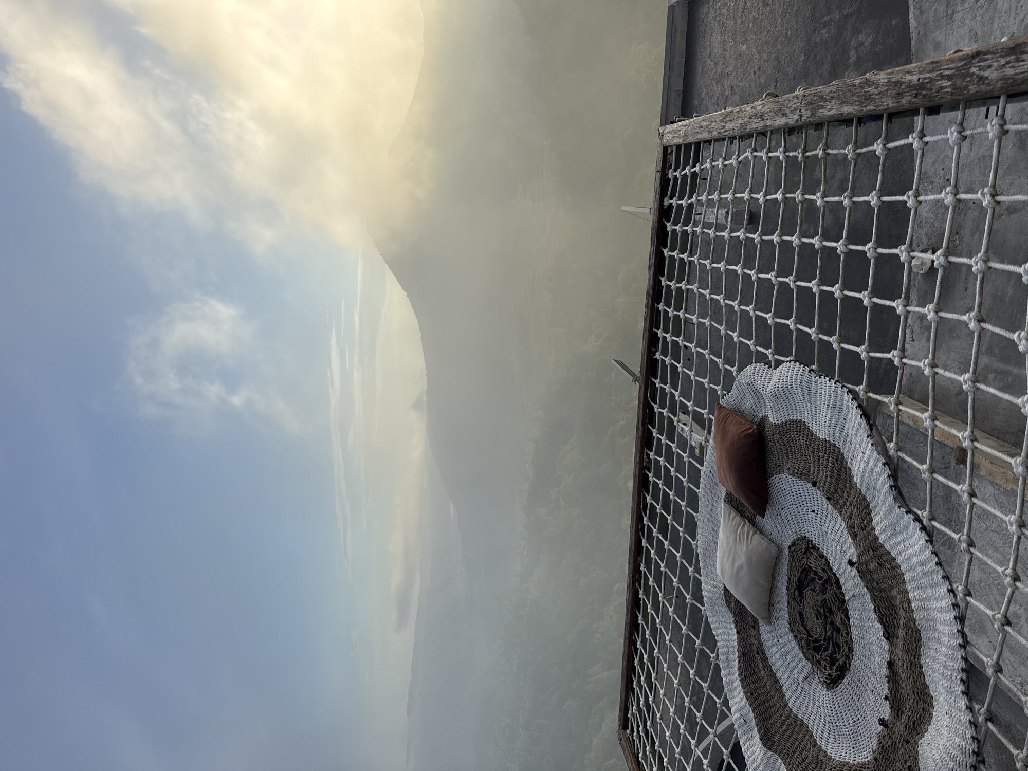 The image depicts a net-like platform with a round striped rug and two pillows, appearing to be an outdoor relaxation area. The location is Kedisan, Indonesia, as suggested by the setting and visual characteristics. The background features a mountainous landscape shrouded in mist and partially obscured by clouds, suggesting a high-altitude location. The sky shows a gradient from blue to yellow, indicative of either sunrise or sunset. The foreground shows the constructed platform, which is designed for sitting or lying, and it has a simple, rustic aesthetic. The rug and pillows provide comfort and visual appeal. The image captures a scenic view and a tranquil setting.