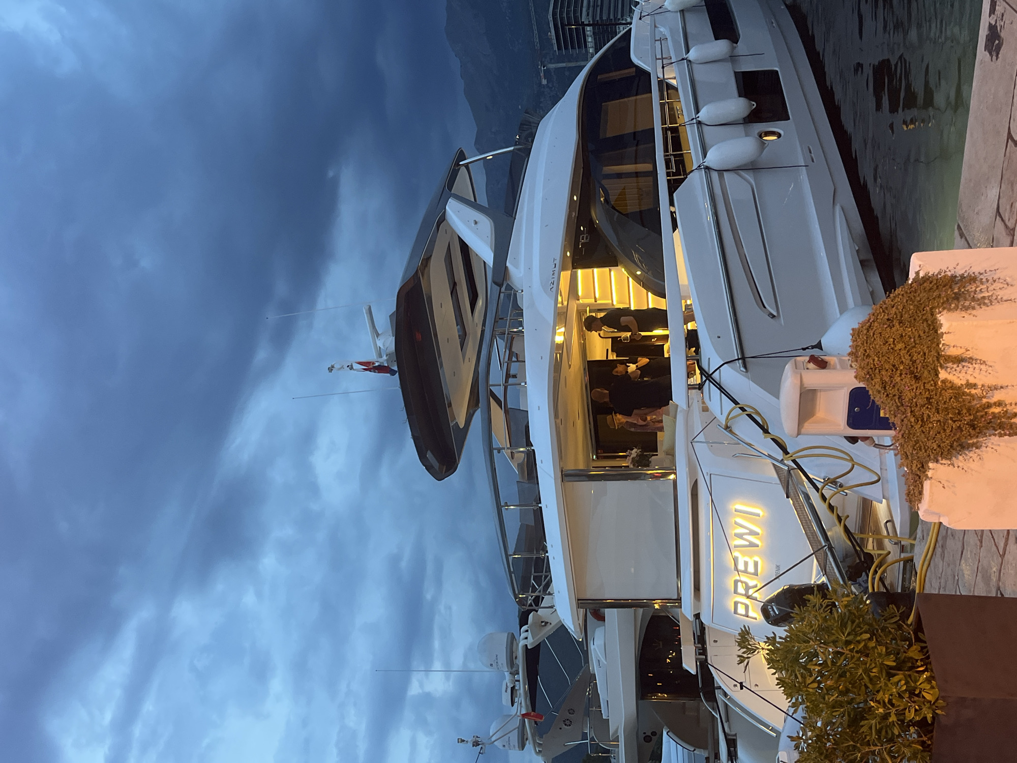 In Budva, Montenegro, a large, white yacht named "PREWI" is docked at dusk. The yacht is illuminated from within, with people visible on deck. The sky is dark and cloudy, with mountains visible in the background. The yacht is equipped with multiple fenders and mooring lines. The scene appears to be a social gathering on the yacht.