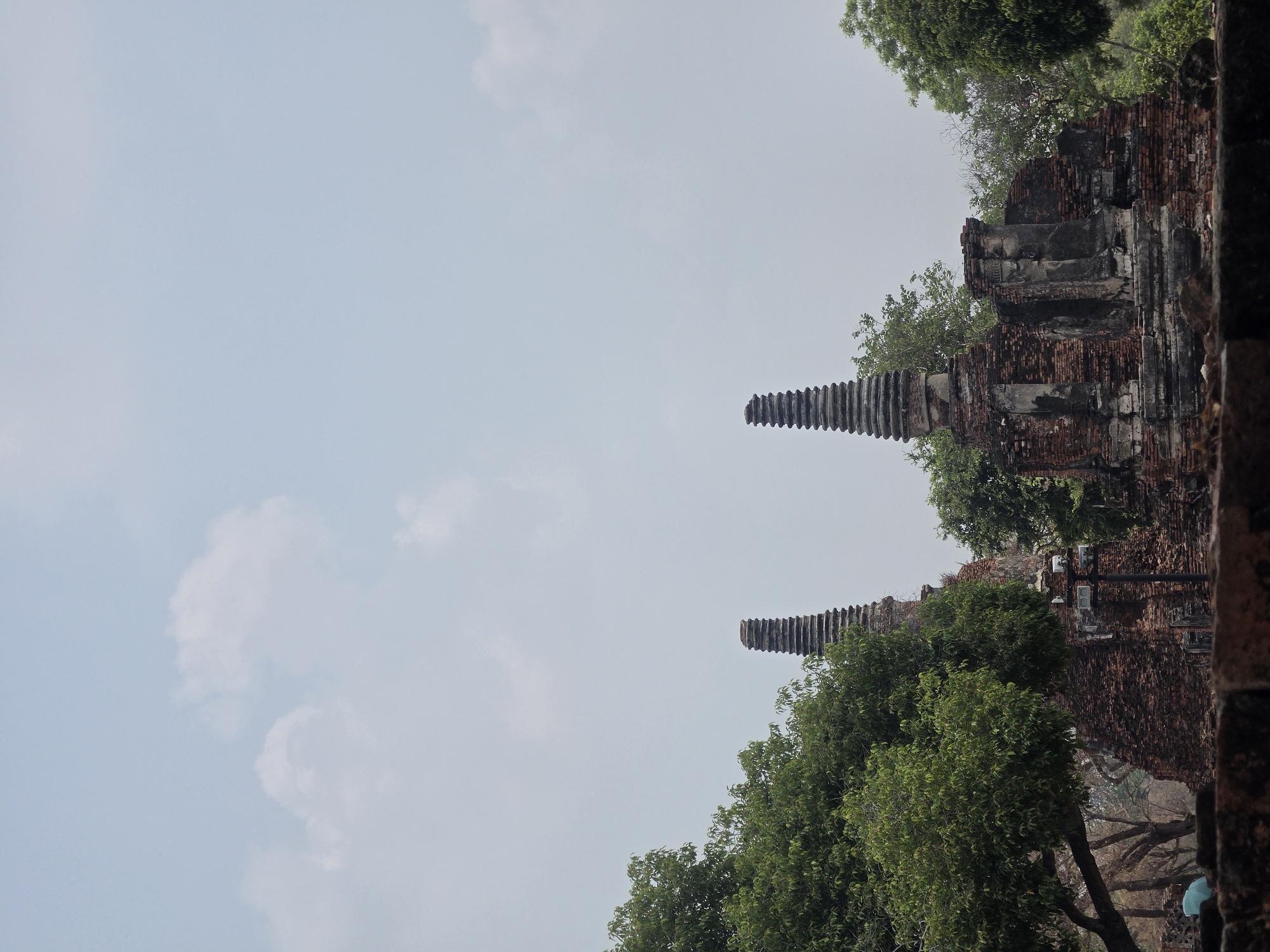 The image depicts an ancient brick structure with tapering towers, partially obscured by trees, against a backdrop of a light blue sky with scattered clouds. The location is likely in the city of Phra Nakhon Si Ayutthaya City Municipality, Thailand, known for its historical ruins. No people are visible. The environment is a historical site, with the structure suggesting a temple or palace ruin.