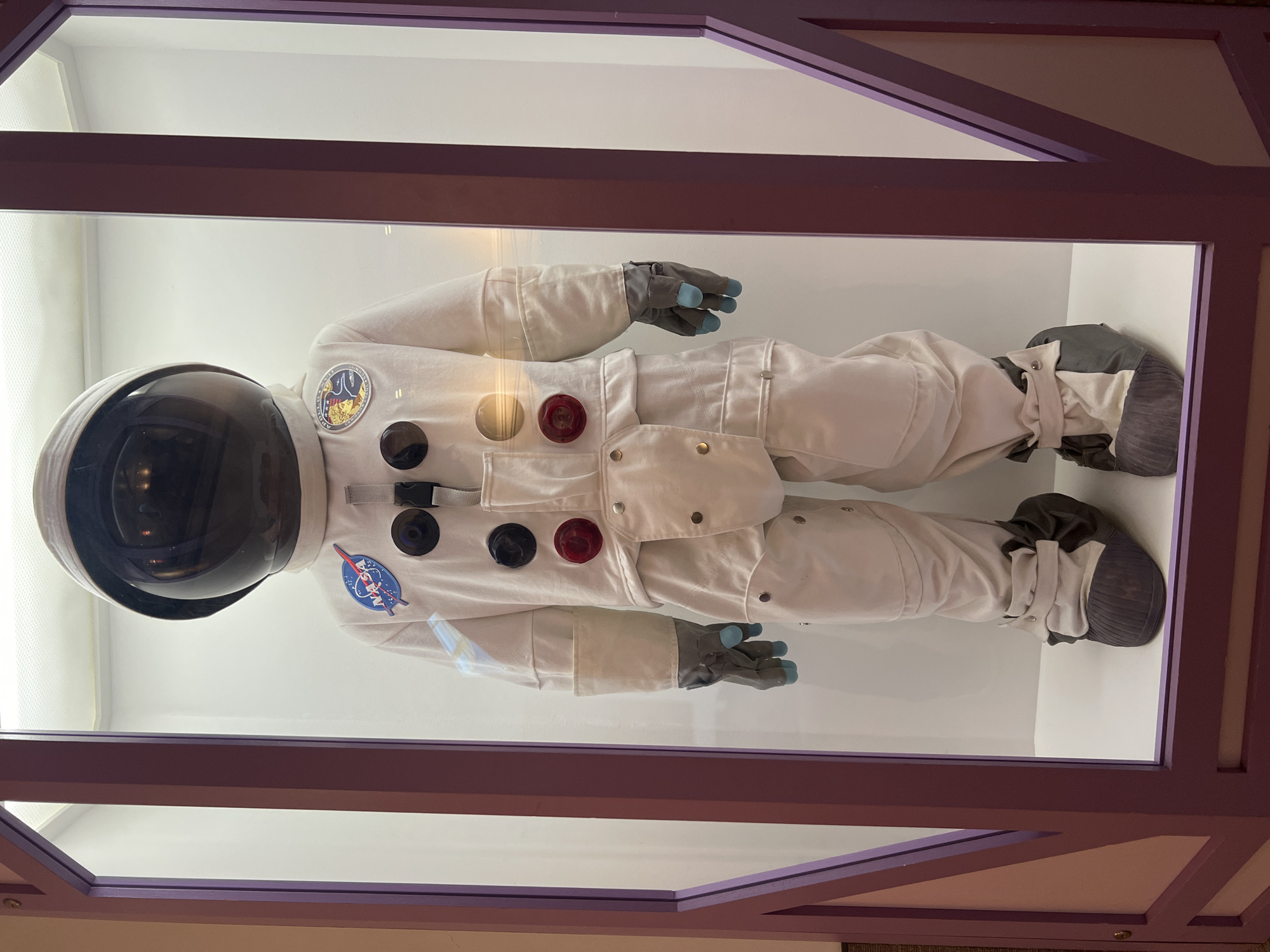 A photograph shows a full-bodied astronaut spacesuit displayed inside a glass case. The spacesuit is predominantly white with various black and red circular features. The spacesuit also has a NASA logo on the chest. The suit includes gray gloves with blue fingertips and gray boots with white straps. The glass case has a purple frame and reflects light. The photograph was taken in Chicago, Illinois, United States.