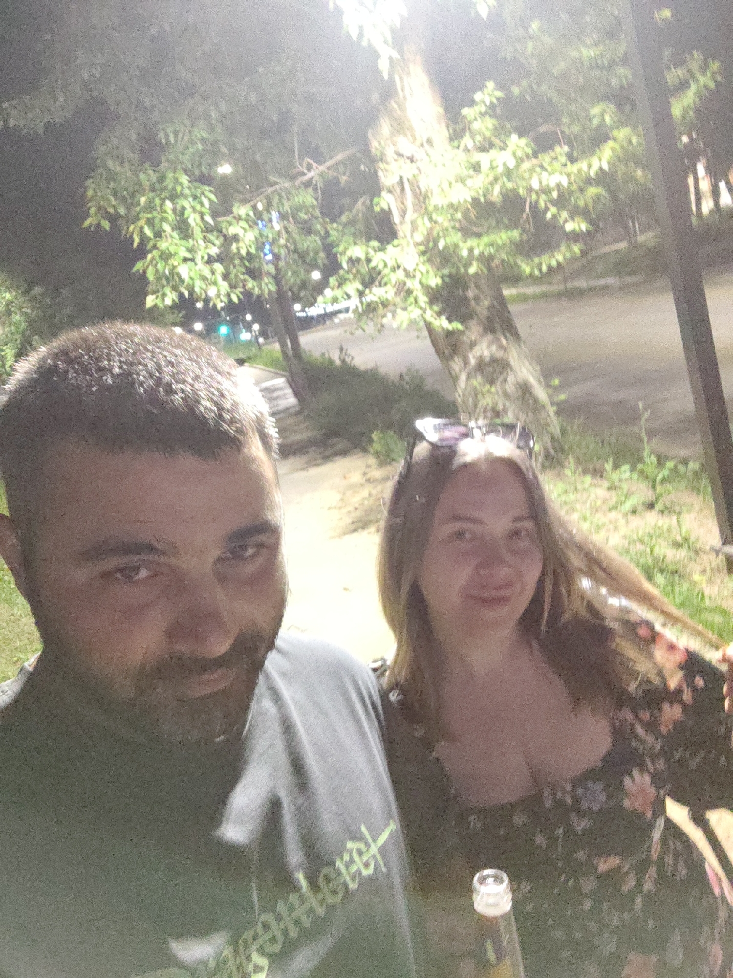 In this selfie taken outdoors at night, a man and a woman are featured. The man, on the left, has short, dark hair, a beard, and is wearing a dark t-shirt with a green design. The woman, on the right, has long, light brown hair and is wearing a dark, floral-patterned dress. Sunglasses are perched on her head. The background shows trees, streetlights, and a paved surface, suggesting a park or a street in a city. The image appears to have been taken in Shimanovsk, Russia, though no specific landmarks are identifiable within the frame.