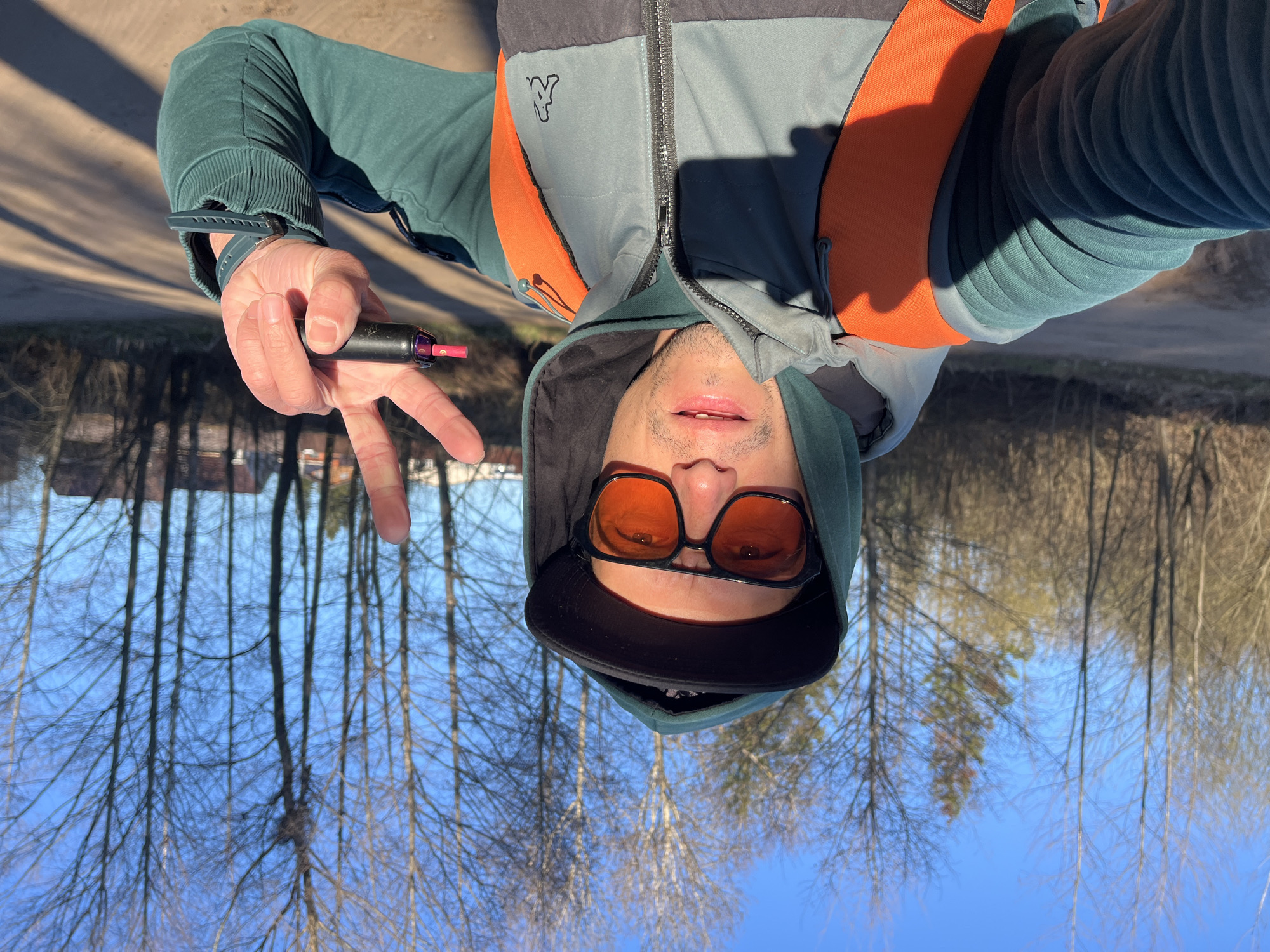 A selfie-style portrait of a man, oriented upside down. The man is looking directly at the camera. He is wearing a dark teal hoodie or sweatshirt with ribbed cuffs, and over it, an orange and gray puffer vest with a black zipper. A small, black stylized 'K' or Kappa logo is visible on the gray section of the vest. He also wears a dark baseball cap under his hoodie and large, orange-tinted sunglasses. His lips are slightly parted.
His right hand (positioned on the viewer's left due to the orientation) is raised, making a peace sign (V-sign). He holds a black vape pen with a prominent red tip between his index and middle fingers. A dark-colored watch or fitness tracker is visible on his left wrist.
The background depicts an outdoor setting under a clear blue sky. Numerous deciduous trees with bare branches are visible, suggesting either late winter or early spring. A dirt path or road is partially visible on the ground. Strong sunlight casts distinct shadows on the man's clothing and the surrounding environment. The scene takes place in Chaikovsky, Russia.