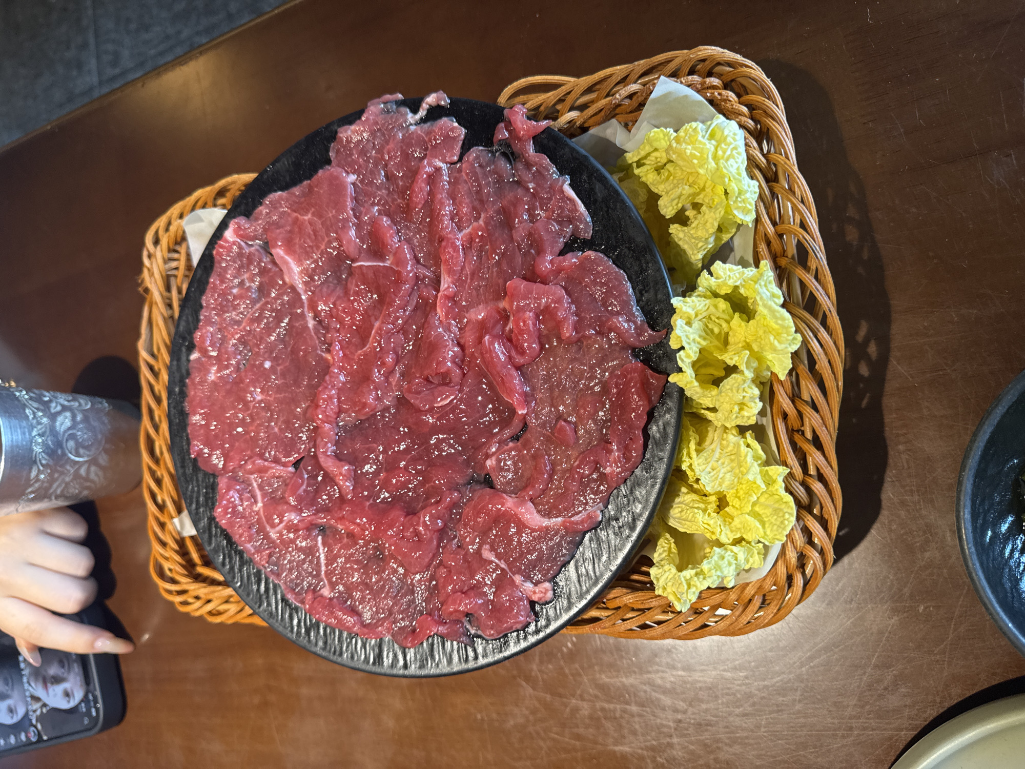 An overhead shot captures a dining scene in Liangjiang, China, centered around raw ingredients prepared for a meal, likely hot pot. A large black circular plate dominates the left side, filled with numerous thin, glistening slices of raw red meat, fanned out in an overlapping arrangement. To its right, a rustic brown wicker basket, lined with white paper, holds bright yellow-green leafy vegetables, resembling napa cabbage.
These food items rest on a dark brown wooden table, which shows visible scratches and wear, characteristic of a busy restaurant or home dining setting. In the lower-left corner, a person's hand with manicured, long fingernails holds a black smartphone. The phone's screen displays an image or filter of a face, along with some indistinct icons. A silver-colored, textured metal cup or container is partially visible above the hand. On the far right, parts of a dark grey bowl and a lighter grey-green plate are also visible at the edge of the frame, suggesting other place settings or serving dishes. The overall lighting is even, indicating an indoor setting. No specific text is clearly decipherable. The scene conveys an anticipation of a fresh, cooked meal.