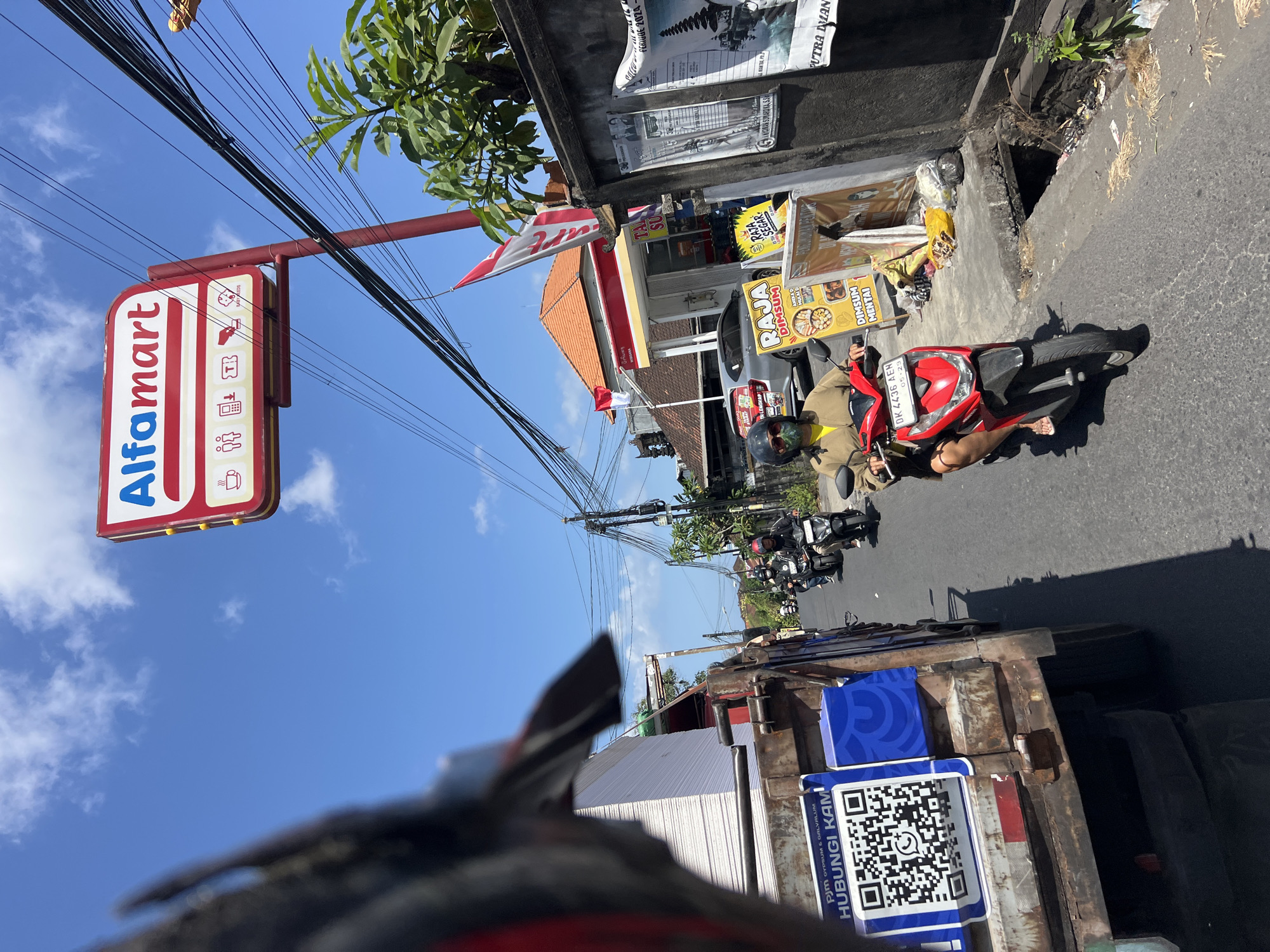 The image captures a street scene in Denpasar, Indonesia, under a clear blue sky with scattered clouds. In the foreground, on the left, a truck is partially visible, loaded with stacks of white boards. A large QR code is affixed to the truck's side, with the text "PJLM GYPSUM & GALVALUM HUBUNGI KAMI" above it. On the right side of the street, there is a red and white Alfamart convenience store. In front of the store, several individuals are seen on motorbikes. One person in a yellow top and dark helmet is riding a red scooter, their face partially obscured by sunglasses and a mask. Further down the street, other motorbikes are visible, with riders wearing helmets. Along the sidewalk, there are posters and signs, including one for "RAJA DIMSUM." The environment appears to be an urban street with electrical wires crisscrossing overhead, and some greenery is present. The overall impression is of a busy, everyday street in a tropical climate.