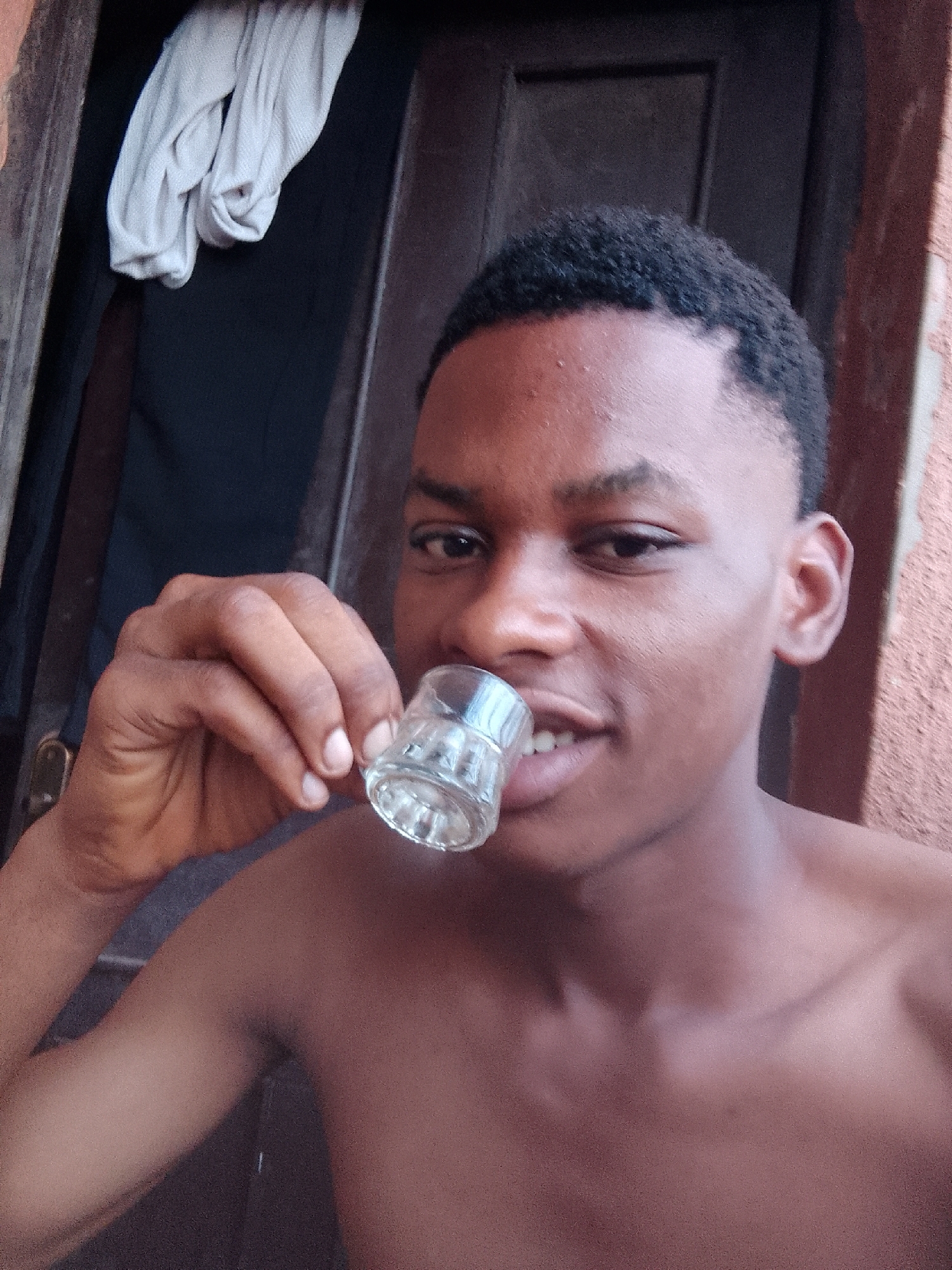 A shirtless young man with short, dark hair is holding a small, empty glass to his lips. He is looking directly at the camera with a slight smile. He appears to be standing in a doorway. A white cloth is hanging in the doorway, and there is a dark brown door frame behind him. The exterior wall on the right is textured and reddish-brown. Given the prompt, the location is likely Nkpor, Nigeria.