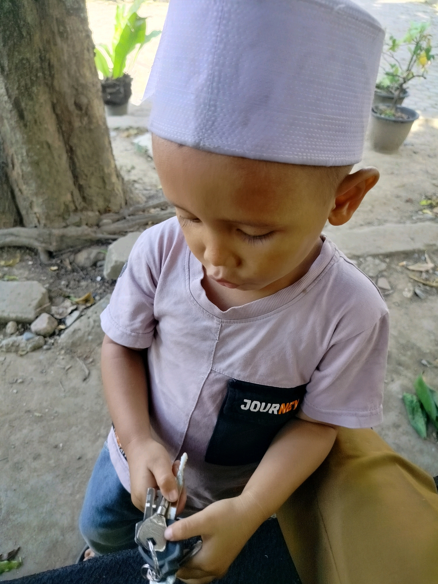 A young boy, wearing a white peci and a light purple t-shirt with a "JOURNEY" patch, sits outdoors on what appears to be a dirt path. He is holding a set of keys in his hands. The background shows a tree trunk, potted plants, and rocky ground, suggesting a rural or garden setting in Purwodadi, Indonesia. The boy is looking down at the keys.