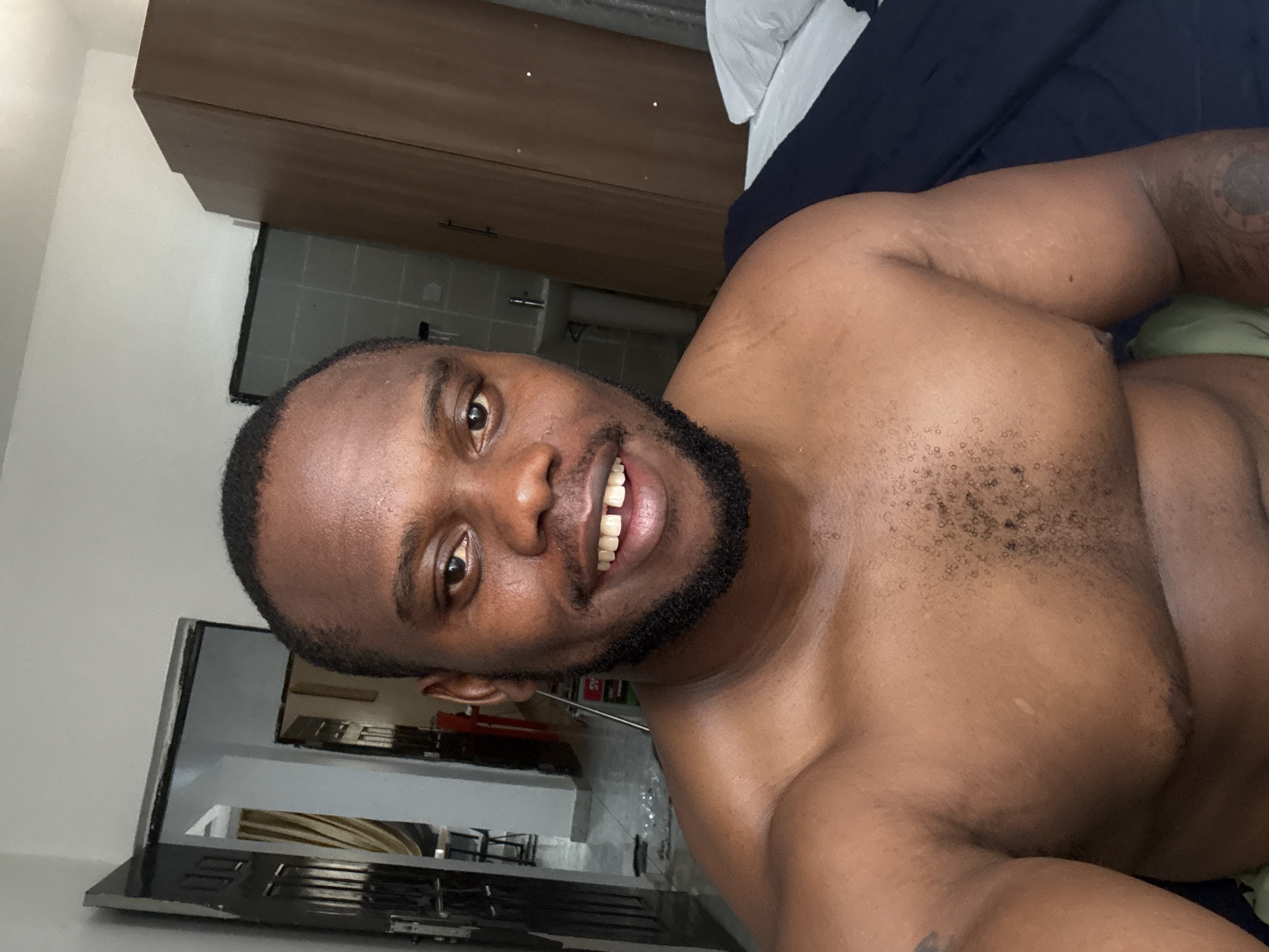 A shirtless man with dark skin, a beard, and closely cropped hair is lying in bed. He is smiling. He appears to be inside a room, possibly a bedroom, with furniture and a glimpse of another room beyond. The image does not provide sufficient information to confirm the location of the photo in Iddo Tudunwada, Nigeria.