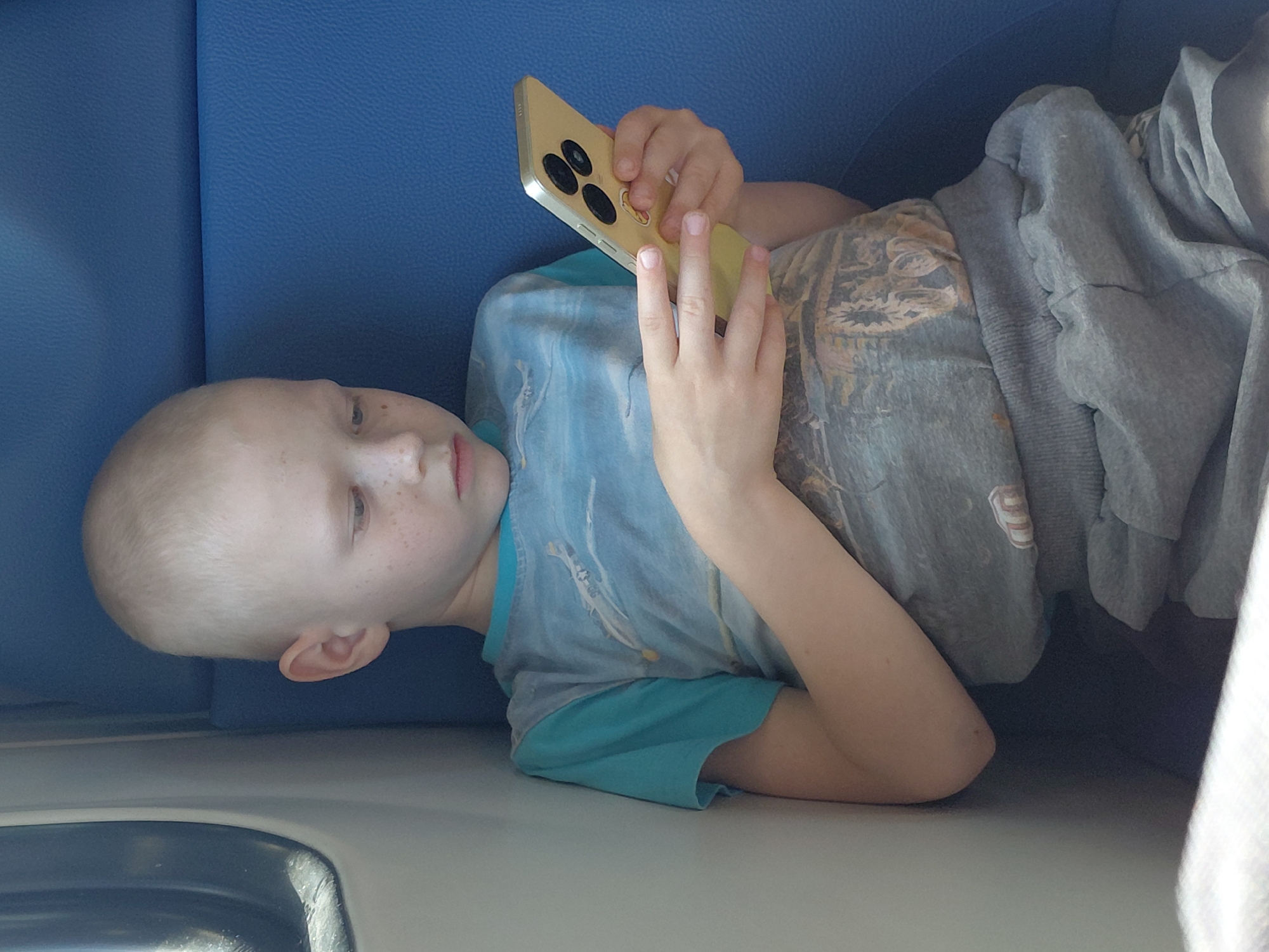 A young, fair-skinned child with short, light hair and freckles is seated, looking down at a yellow smartphone held in both hands. The child is wearing a blue t-shirt with a graphic print and grey sweatpants. The setting appears to be a vehicle interior, with a blue seat and a window visible. There are no specific landmarks or indicators that place this image in Barmashevo, Russia. The child's posture and engagement with the phone suggest a moment of personal use.