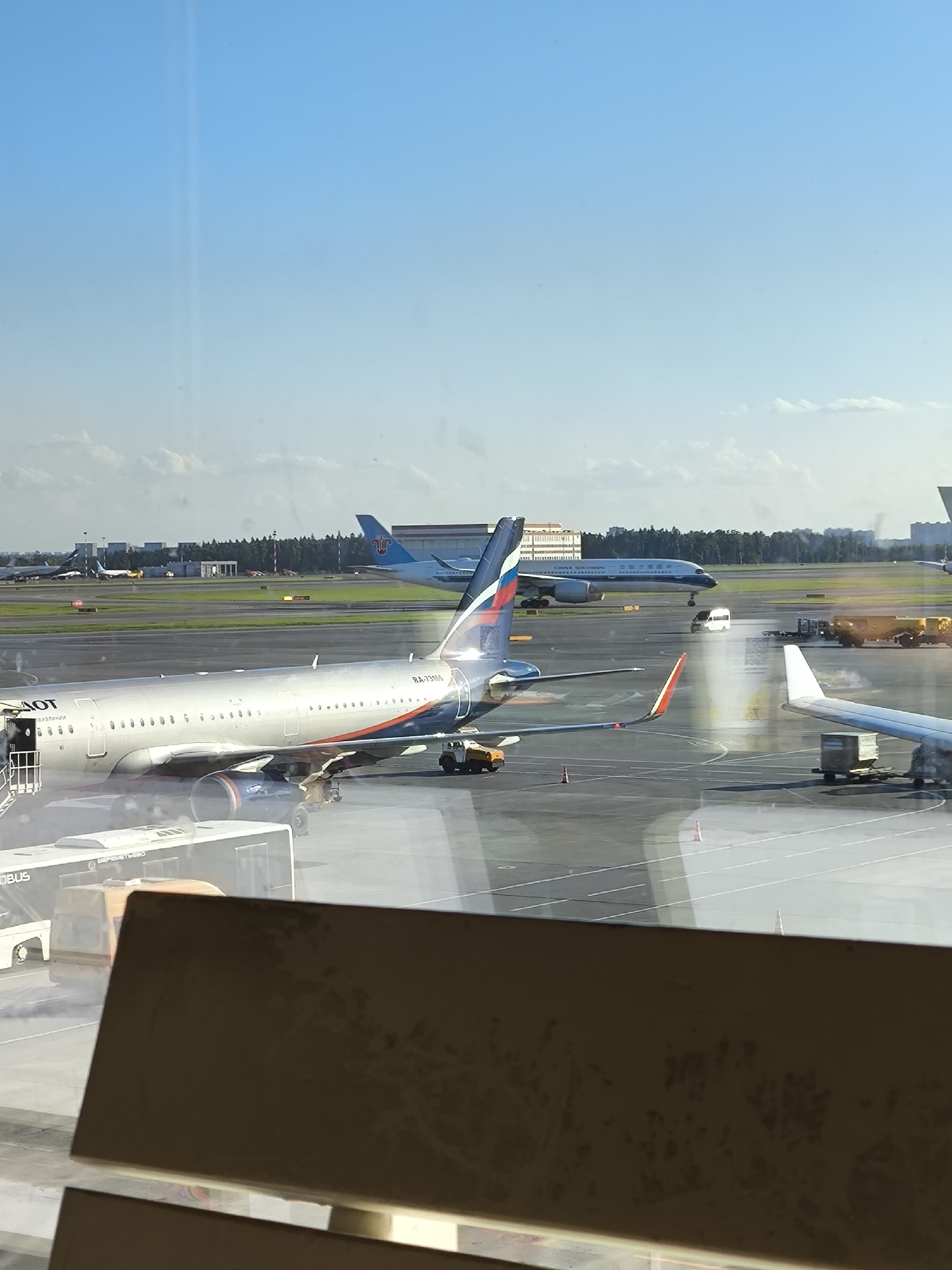 The image presents a sunlit view of an airport tarmac through a window, featuring multiple aircraft and ground support vehicles. In the left-center foreground, a silver-colored Aeroflot single-aisle aircraft is parked, displaying a blue and red livery on its tail with the registration "RA-73186" visible. The text "AOT" is partially visible on its fuselage. A mobile boarding stair is attached to the front-left door of this aircraft, and its engines are dark blue/grey.
Further in the background to the left, a white China Southern Airlines wide-body aircraft is visible, featuring a blue tail with its logo and "CHINA SOUTHERN" text in both English and Chinese characters on its fuselage. On the right side of the tarmac, a portion of another white aircraft's wing and fuselage is visible.
Ground support equipment is active around the aircraft. A white passenger bus with "MEREPOMETBO" written on its side is partially visible in the lower left foreground, near the Aeroflot plane. A yellow fuel truck and a smaller white van are seen further right on the tarmac, with some exhaust smoke or vapor visible near the rear of the Aeroflot aircraft. The tarmac is grey with white painted markings and yellow lines, and several orange traffic cones are dispersed across it.
The background shows airport infrastructure, including various buildings and a line of green trees under a clear, bright blue sky with a few scattered white clouds along the horizon. A faint vertical light streak, indicative of a reflection on the viewing window, is present in the upper central sky. The bottom quarter of the image is obscured by a dark, textured, horizontal brown structure, likely a window frame or part of a piece of furniture, showing some light reflections.
