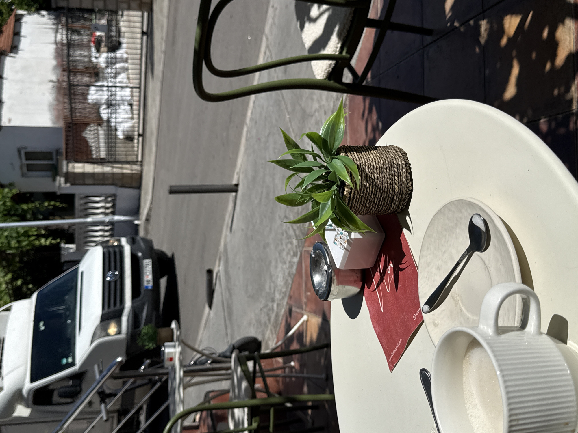 A table at an outdoor cafe in Budva, Montenegro, features a white ceramic mug filled with a frothy beverage, a white saucer with a silver spoon resting on it, a small potted plant in a woven basket, and a metal ashtray. The table is set against a backdrop of a street scene, with a white van parked on the left and a building with a metal gate and white debris visible on the right. Shadows from trees dapple the ground, indicating a sunny day. The overall scene suggests a moment of relaxation at a cafe.