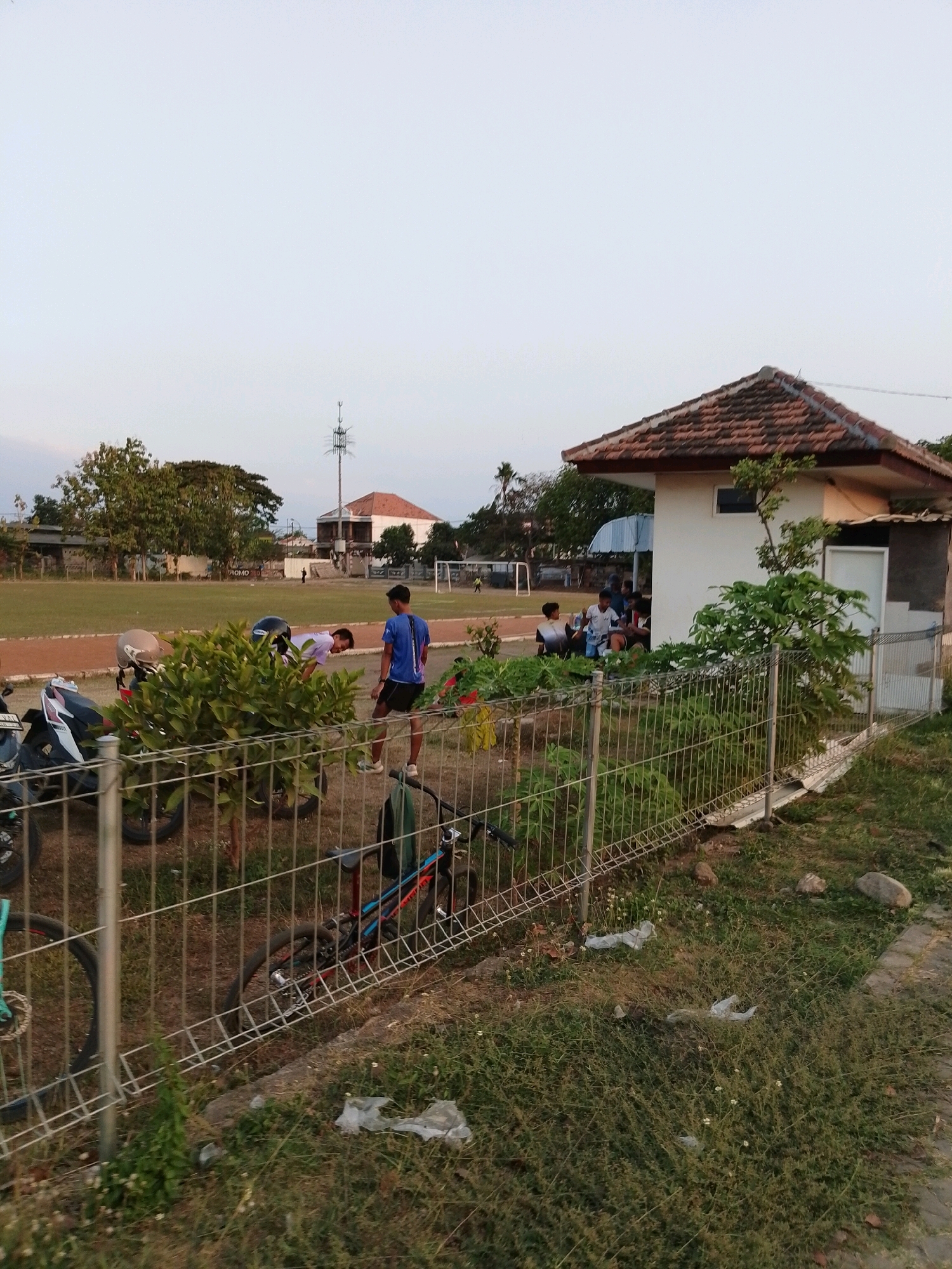The image depicts an outdoor scene near a sports field in Pasuruan, Indonesia. In the foreground, a wire fence separates the viewer from the field. Several motorcycles and bicycles are parked behind the fence. To the right, a small white building with a tiled roof stands next to a lush green bush.
Beyond the fence, a grassy field stretches out, possibly a sports field, with a running track visible. In the mid-ground, several individuals are present on the field. Some appear to be playing or engaging in physical activity, while others are gathered near the building. A young man in a blue t-shirt and black shorts is standing, looking towards the field. Another person in a white t-shirt is bending over near a bush. In the distance, a telecommunications tower rises against a pale sky. The environment suggests a casual outdoor gathering, likely a recreational area.