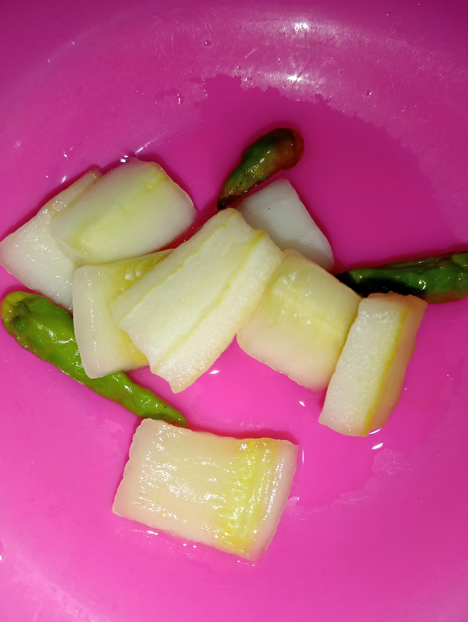 A bright pink bowl contains several pieces of white vegetable, likely daikon radish, cut into thick rectangular shapes, along with three green chili peppers. The vegetables and peppers appear to be submerged in a light liquid, possibly water or brine, which creates reflections on the smooth surface of the bowl. The scene is captured from a top-down perspective. The location is described as Loa Janan Ulu, Indonesia.