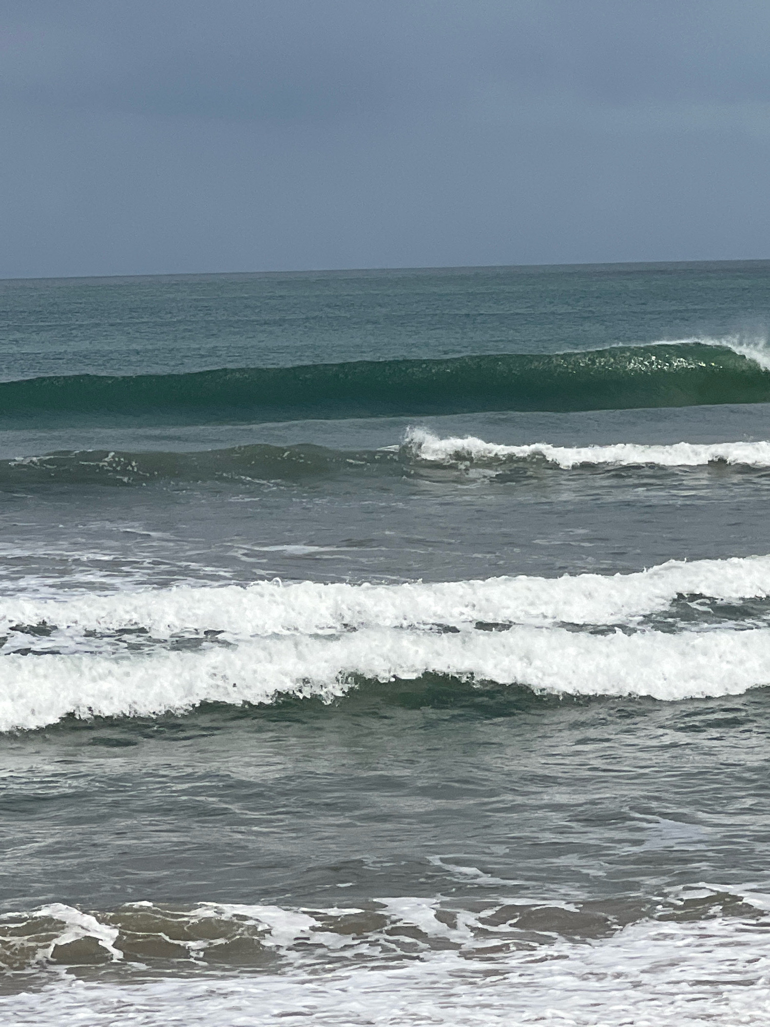 The image captures a series of waves breaking on a beach under a cloudy sky. The water is a deep green, transitioning to white foam as the waves crest and crash. In the distance, a larger, more powerful wave is visible, appearing to be in the process of forming or breaking. The sky is overcast with a muted, gray hue, suggesting a subdued or possibly stormy weather condition. The foreground shows the choppy, white-capped water nearing the shore, with hints of darker sand or seabed beneath the surface. The scene is devoid of any people or structures, focusing solely on the natural phenomenon of the ocean. This image depicts a typical seascape found along the coast of Kuta, Indonesia, known for its surf breaks and energetic ocean currents.
