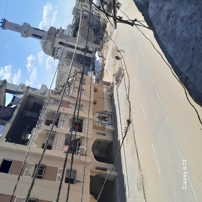 The image depicts a severely damaged urban landscape in the city of Khan Yunis, Palestinian Territory, under a sky with scattered white clouds. The foreground is partially obscured by numerous dark utility cables spanning horizontally. To the left, a multi-story, light-colored residential building shows extensive structural damage on its upper four floors, with large sections of walls missing, exposing internal spaces and debris. Its lower floors appear more intact, featuring balconies, some with hanging fabrics, and a ground-level commercial unit with arched entryways and a storefront sign. Adjacent to this building and extending to the right, a mosque with a domed roof and a minaret is visible, exhibiting significant damage to its upper sections and surrounding architectural elements. Large piles of rubble, concrete fragments, and twisted metal debris are scattered on the ground surrounding the damaged structures, with some areas partially covered by white tarpaulins. A paved street with faint white lane markings traverses the mid-ground, bordered by a concrete sidewalk. A single individual on a dark motorcycle is observed traveling on this street, moving away from the viewer towards the right side of the frame. The overall environment indicates widespread destruction.