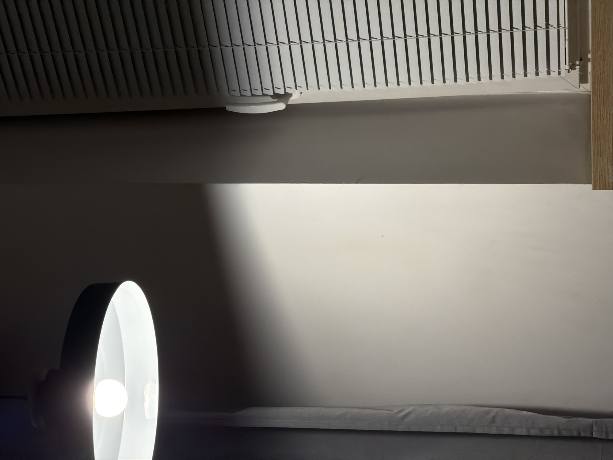 The image depicts an interior scene with a light fixture overhead casting a bright light onto a wall. To the right of the wall, a window with closed blinds is visible. A grey curtain hangs on the left side of the image. The overall lighting suggests an indoor environment, possibly a room in a residential or commercial building. The exact location in Sosnowiec, Poland, cannot be determined from the visual information provided. There are no people, actions, or interactions depicted in the image.