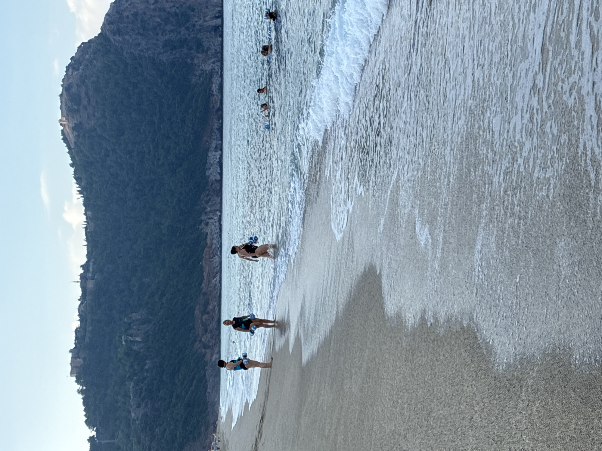 The image depicts a scene at the beach in Alanya, Turkey, with the Alanya Castle visible on the mountain in the background. In the foreground, several women are standing in the shallow water and on the wet sand, holding what appear to be flotation devices or weights for water aerobics. In the distance, several people are swimming or playing in the sea. The sky is partly cloudy, and the overall atmosphere is that of a recreational day at the beach.