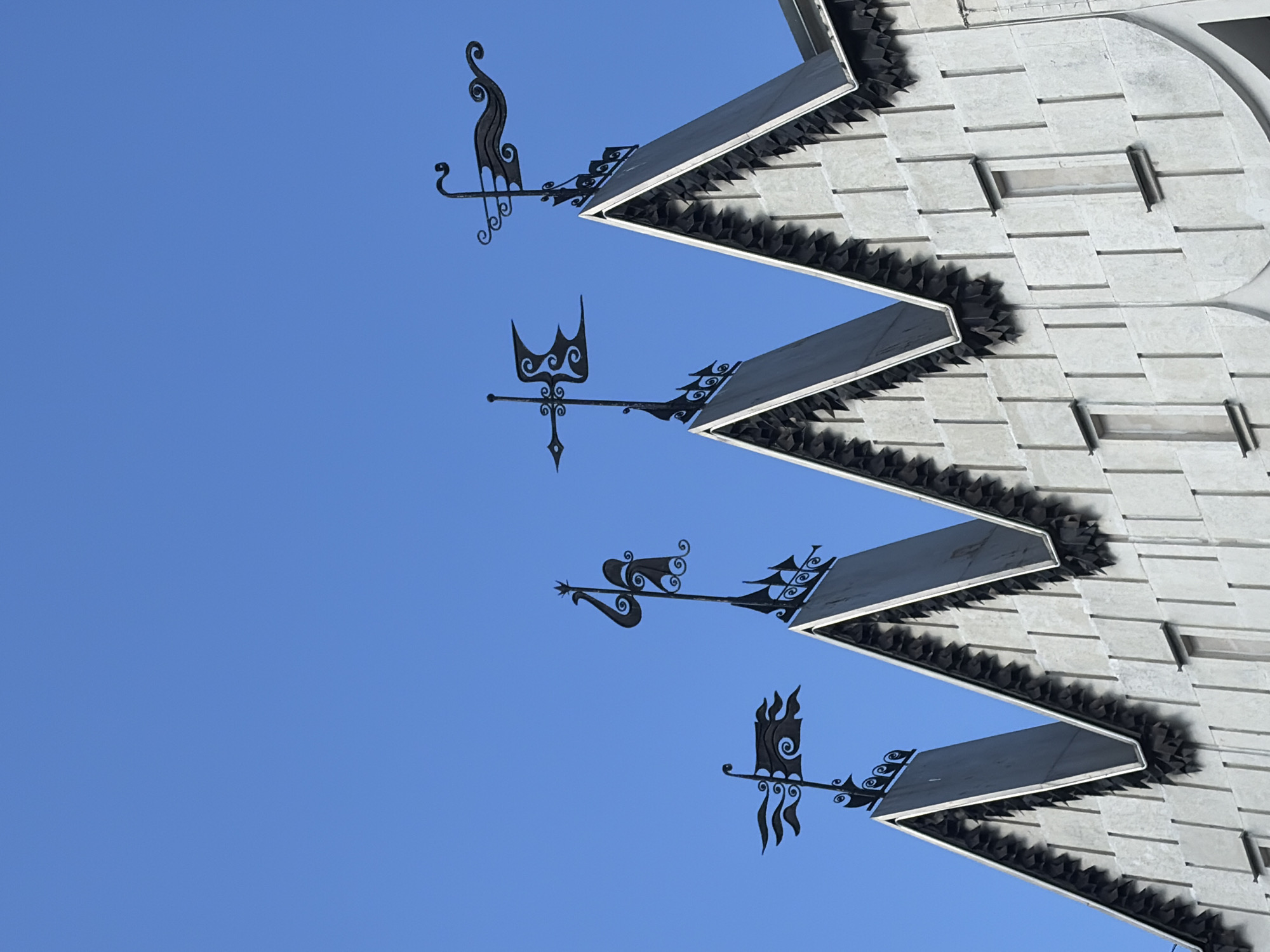 The image shows a portion of a building facade with a clear blue sky in the background. The building has a unique architectural style with sharp, triangular roof sections and a textured concrete exterior. Each triangular section is topped with an ornate black metal weather vane. There are at least four visible weather vanes, each with a distinct design. The facade features several narrow, vertical windows with dark frames. The overall impression is of a modern or stylized building with decorative elements on its roof. There is no information in the image to determine its specific location in Nizhny Novgorod, Russia.