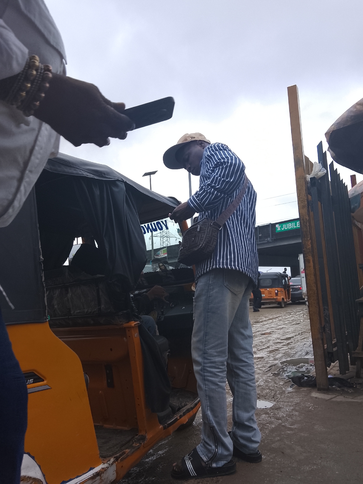 In Lekki, Nigeria, under an overcast sky, a man in a blue and white striped shirt, light blue jeans, and black sandals stands beside an orange auto-rickshaw. He is looking down at his phone, which he holds in his right hand. A brown messenger bag with a patterned strap hangs across his chest. His left hand, adorned with multiple beaded bracelets, is also near the phone. To his left, a portion of another person's arm, wearing a white shirt and bracelets, is visible, holding a dark object, possibly another phone. The auto-rickshaw has a black canvas top. The ground is wet and muddy, suggesting recent rain. In the background, a yellow pole stands near a black metal fence. Further back, a road with traffic, including other auto-rickshaws and cars, is visible, along with an overpass with a green sign that reads "JUBILEE." The scene depicts a moment of everyday urban life in Lekki.