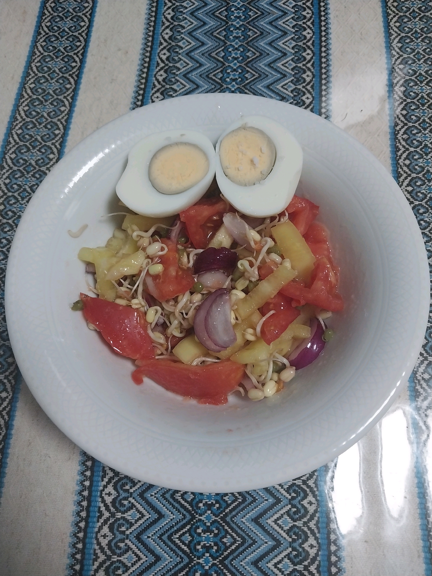 A white ceramic plate containing a salad and two halves of a hard-boiled egg sits on a table covered with a striped cloth. The salad is a mix of chopped tomatoes, sprouts, and possibly some diced yellow bell pepper or pineapple, with slices of red onion mixed in. The two halves of the hard-boiled egg, with their yolks exposed, are placed on top of the salad, resembling eyes. The background cloth has a pattern of blue and white geometric shapes. The lighting suggests an indoor setting, possibly a kitchen or dining area. There is no indication of the location being in Dnipro, Ukraine.