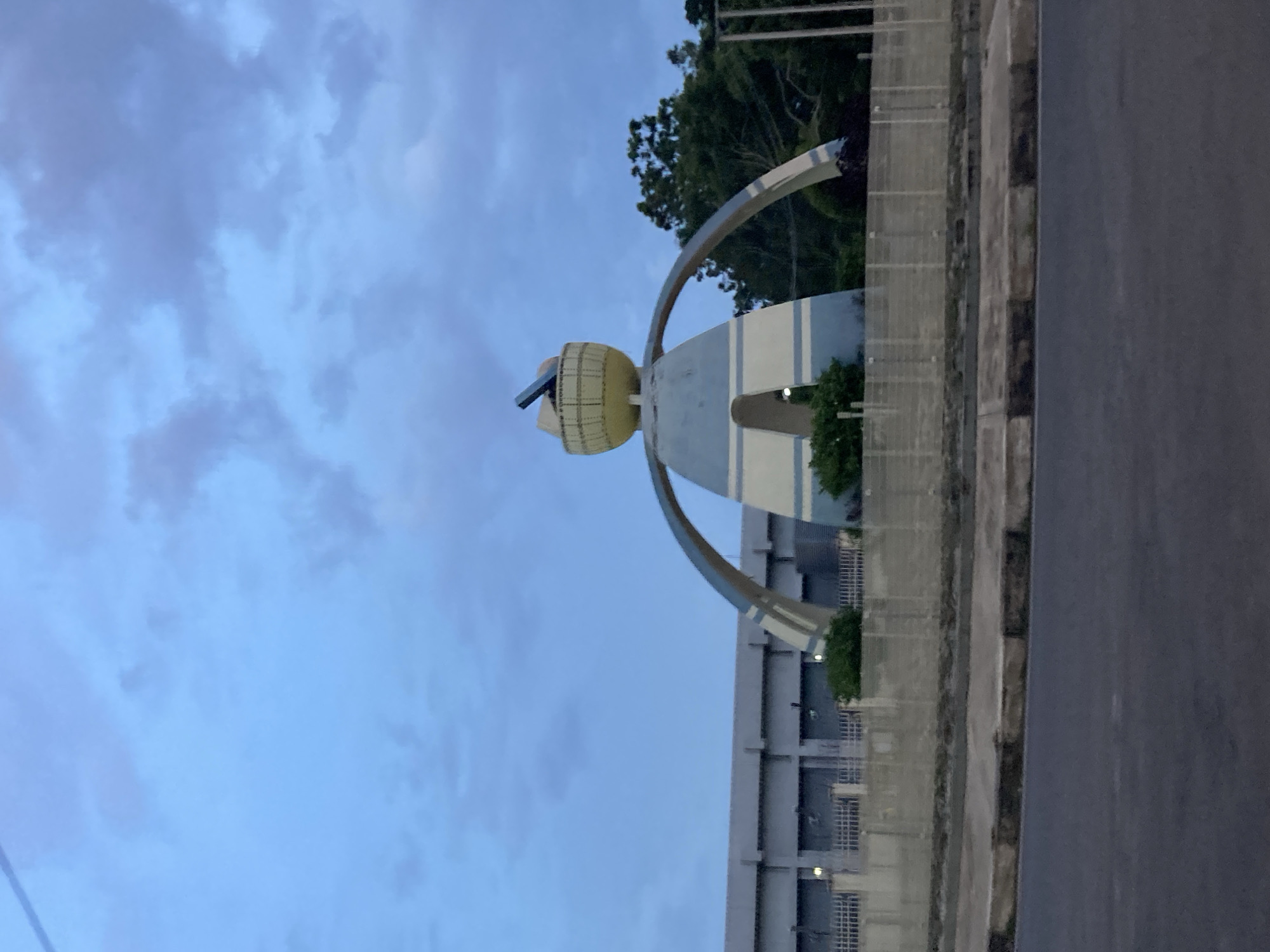 The image shows the "Endurance" statue in Jos, Nigeria. The statue is a large, abstract sculpture made of concrete and metal, depicting a human-like figure with outstretched arms forming arches. The statue is painted in shades of blue and white, with a yellow sphere at the top that holds a dark, angled object. The statue is surrounded by greenery and a fence. In the background, there is a long, grey building with a series of large doors or openings. The sky is cloudy and a light blue, suggesting it is either early morning or late evening. A road is visible in the foreground. The overall scene conveys a sense of public art and urban landscape.