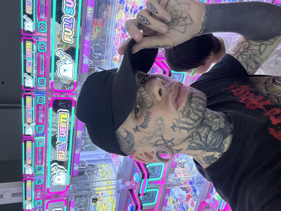 A heavily tattooed individual wearing a black t-shirt and black baseball cap stands in front of arcade claw machines labeled "FULL SUERTE". The person's neck and face are covered in intricate tattoos, including script and floral designs. Their arms also feature dense, dark tattoos. A second person with short dark hair is partially visible in the background. The claw machines are brightly lit with pink and teal neon accents.