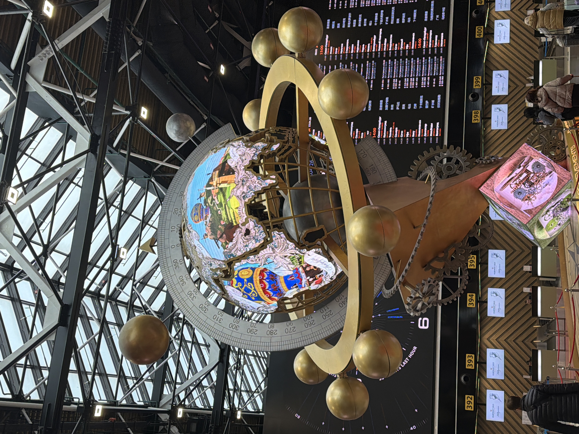 The image depicts an elaborate installation, likely an art piece or an interactive exhibit, inside a building in an unknown city, unknown. The central feature is a globe-like structure decorated with images and surrounded by golden spheres. A metallic arm with gears extends from the globe towards a large digital display that shows various information, possibly time zones or data related to different cities.
Adjacent to the display is a row of small screens or signs with images and possibly text. Several people are visible in the background, some standing and observing the installation, and others walking past, suggesting the exhibit is located in a public area. The architecture of the building features a black metal frame and the presence of lights.