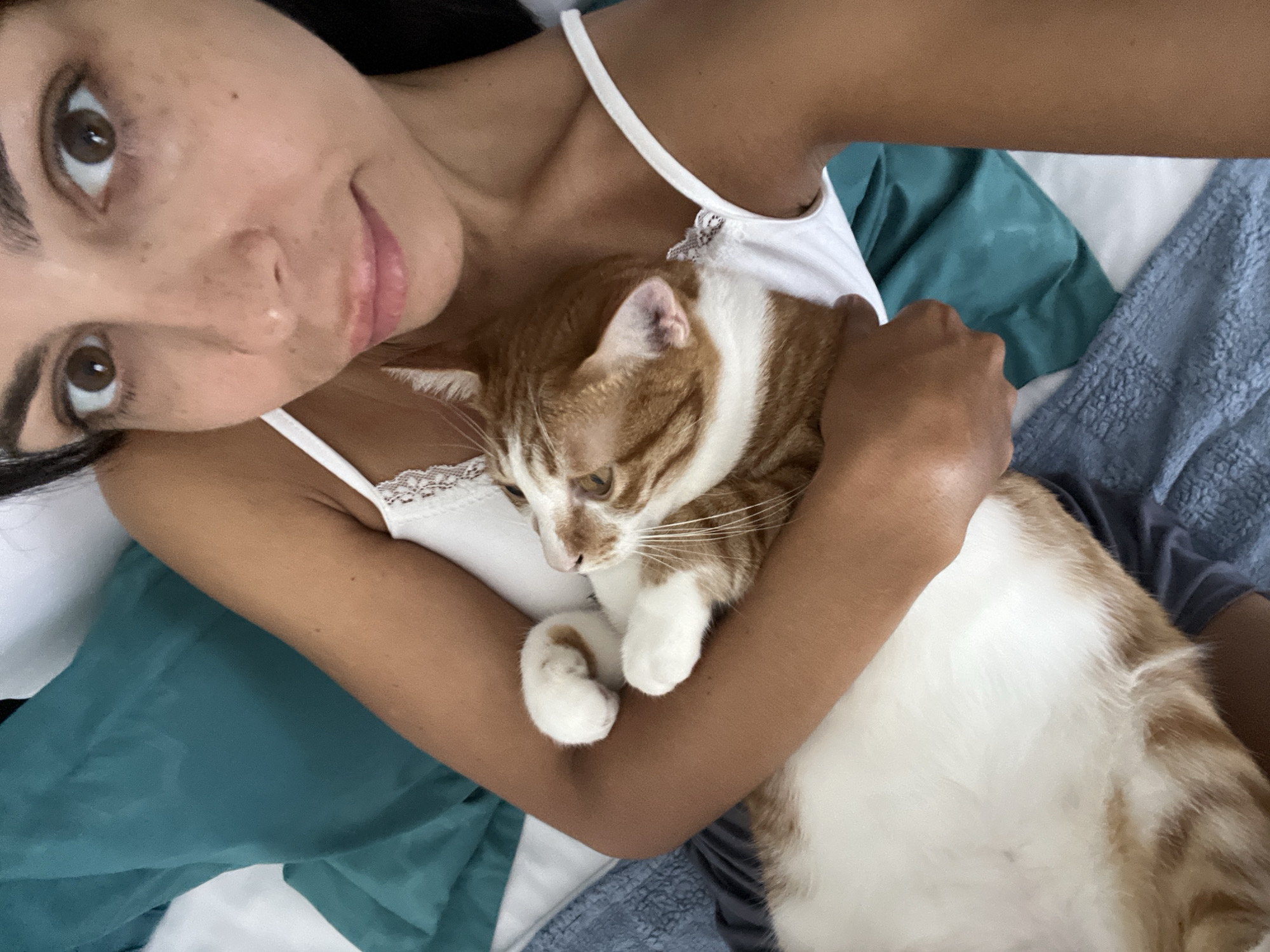 A woman is lying down holding an orange and white tabby cat in her arms. The woman is fair-skinned with brown eyes, dark hair, and pink lips. She is wearing a white tank top with lace detailing. Her arms are wrapped around the cat, which has its eyes open and is looking down. The background consists of bedding in various shades of blue and white. The location is Unknown, Indonesia. The city is unknown.