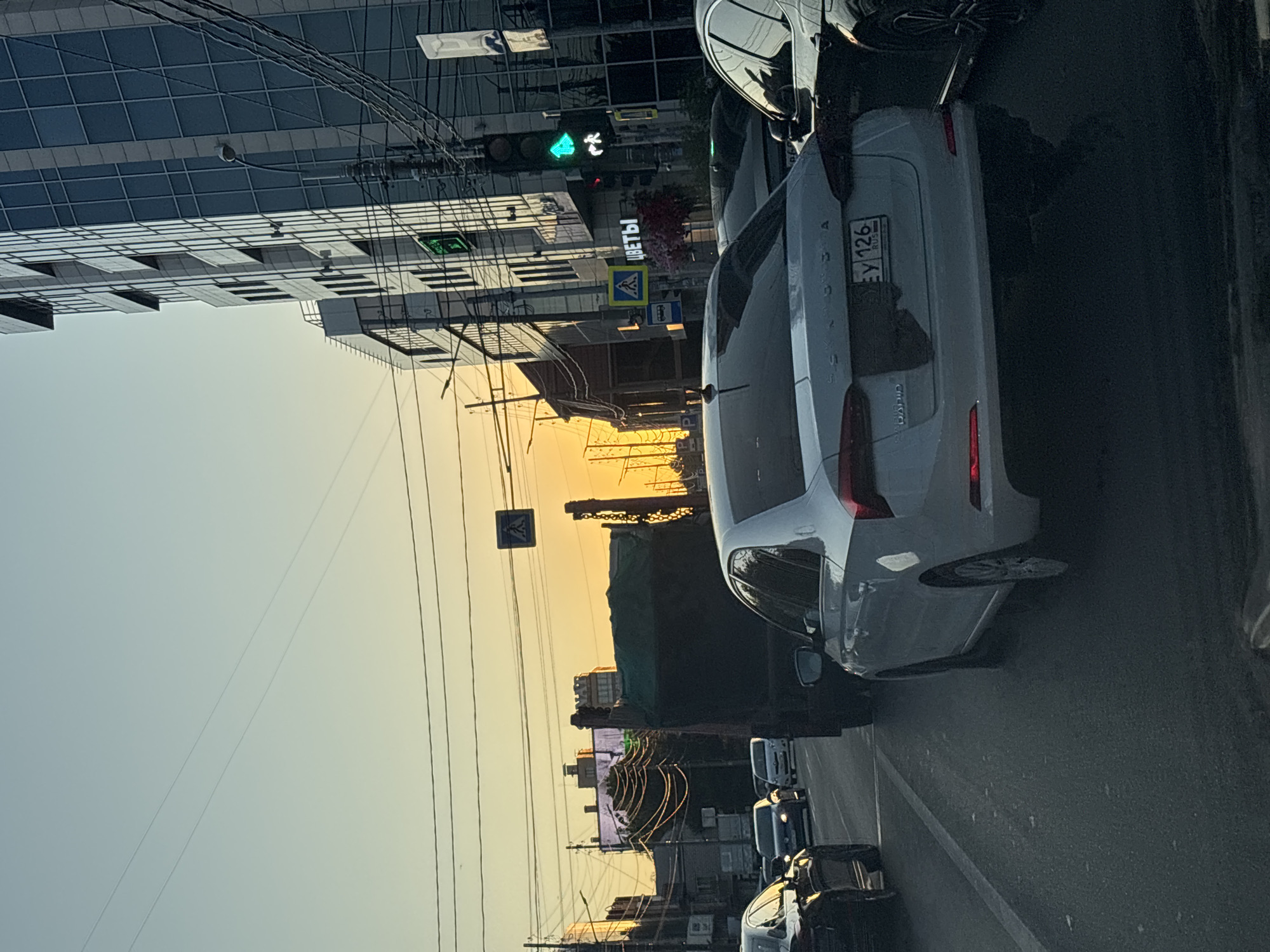 The image captures a street scene in Krasnodar, Russia, during what appears to be late afternoon or early evening, indicated by the warm, low-angled sunlight and the soft, gradient sky. The perspective is from within a vehicle, looking forward at a traffic jam.
Dominating the foreground is the rear of a white sedan, a Skoda, with its brake lights subtly illuminated. Its license plate, partially obscured, reads "EY 126 RUS." To the right and slightly behind the Skoda is a dark-colored car, possibly a luxury sedan, its front portion visible. To the left, the rear of a dark sedan is also visible.
Further ahead, a large, dark-colored truck, possibly a dump truck with a tarp covering its load, is stopped in traffic. Beyond the truck, a line of other vehicles continues down the road. The road itself is asphalt with visible lane markings.
The urban environment is evident. On the right side of the street, a modern, multi-story glass-facade building stands prominently. Electrical wires and poles are a significant feature, crisscrossing the sky, typical of Russian urban infrastructure. A traffic light with a green pedestrian signal and a green arrow is visible, indicating traffic flow. Across the street, a building with ornate architectural details and signage reading "ЦВЕТЫ" (Flowers) is partially obscured by the truck and overhead wires. A pedestrian crossing sign is also visible. In the distance, other buildings and streetlights can be seen. The overall impression is of a busy city intersection during a transitional time of day.