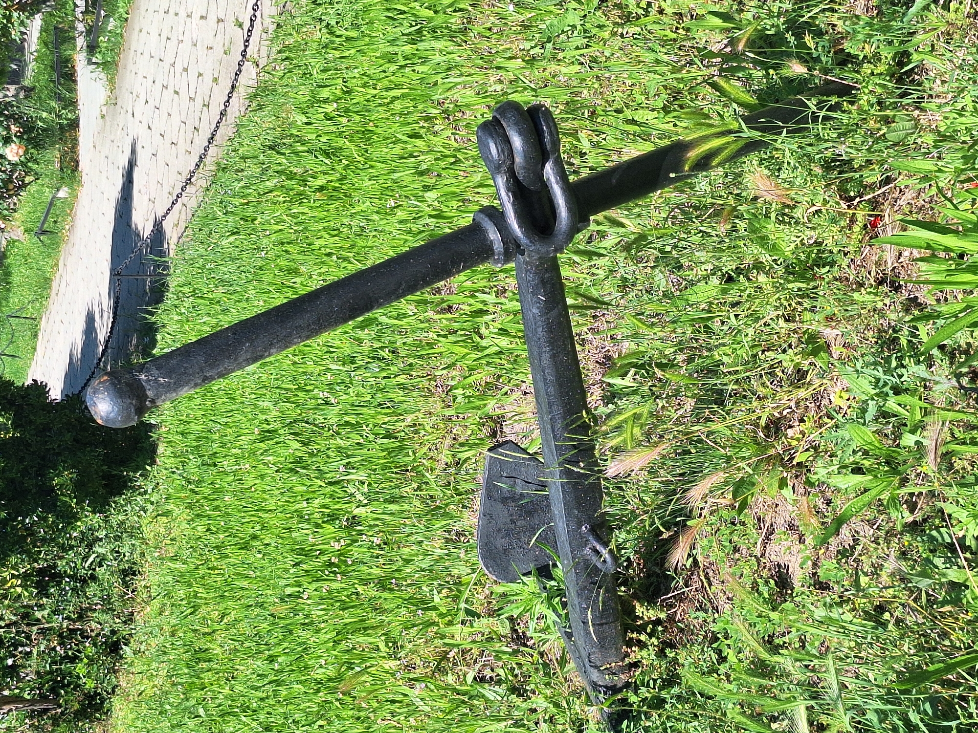 The image shows a close-up of a black anchor lying on a patch of vibrant green grass. The anchor is made of dark metal and appears to be quite old and weathered. One fluke is clearly visible, along with the shank and the shackle at the top. The anchor is positioned diagonally across the frame, with one end resting in the grass and the other extending towards the top left.
To the left of the anchor, a paved pathway made of light-colored stones is visible, bordered by a dark metal chain. The pathway casts a shadow onto the grass, indicating that the sun is shining. The scene is bathed in bright, natural light, suggesting it is daytime. The grass is lush and appears well-maintained, with some taller blades and seed heads interspersed.
In the background, more greenery is visible, possibly bushes or trees, and some other less distinct elements that are out of focus. The overall setting appears to be an outdoor area, possibly a park, garden, or public space. There is no visible text in the image. The anchor's placement on the grass suggests it is a decorative element or perhaps a historical artifact on display, rather than actively being used. The image has a natural, outdoor feel, with the focus sharply on the anchor and its immediate surroundings. The composition highlights the texture and form of the anchor against the softness of the grass.