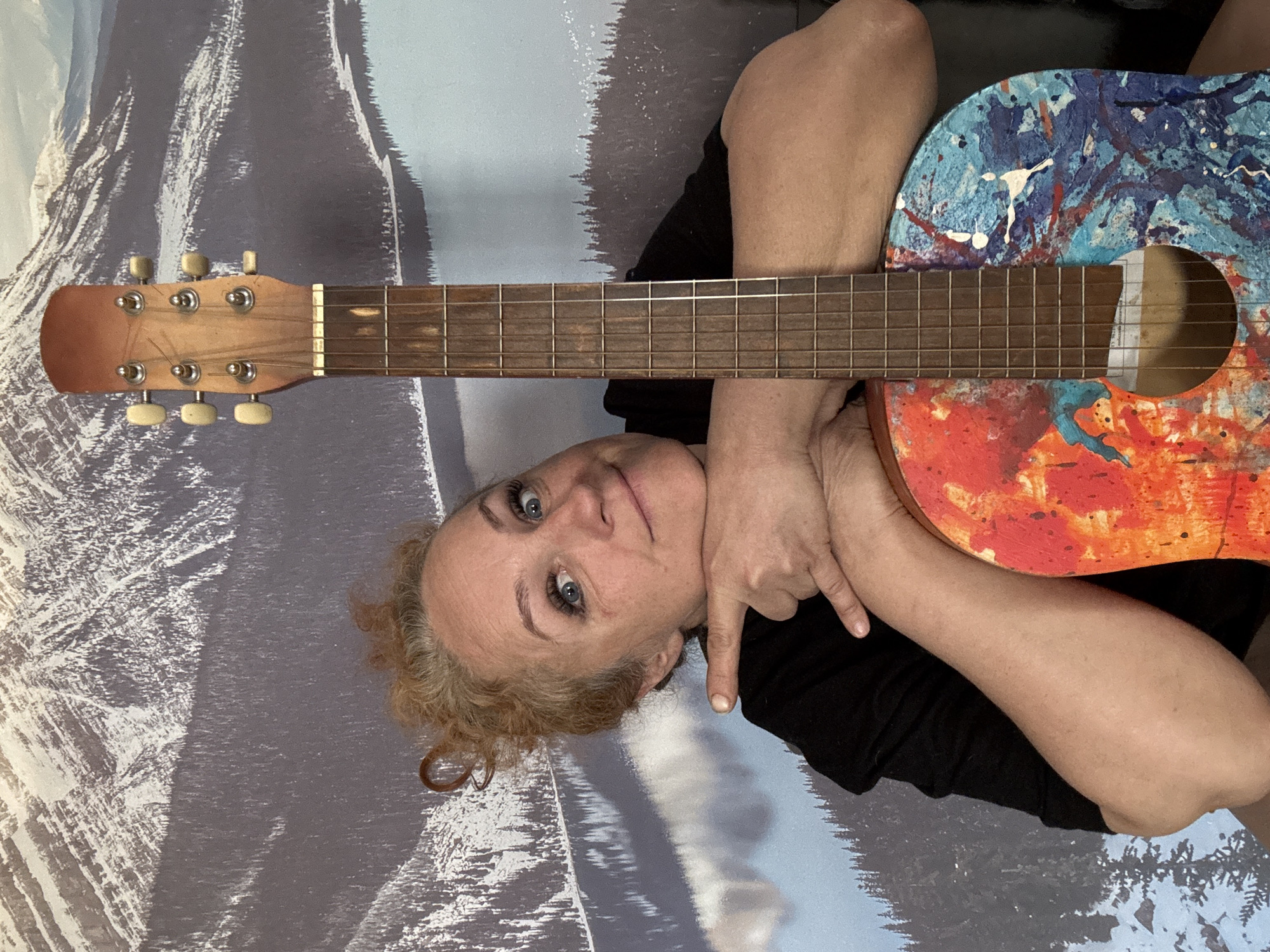 A woman with fair skin and curly blonde hair is positioned with her head to the left, against a background that appears to be a mural depicting a snowy, mountainous landscape. She is wearing a black top. Her right arm is across her body, holding a brightly colored guitar with a blue and orange painted design on it. She is pointing down with her right index finger. The headstock of the guitar is at the upper left of the frame and the body is on the right. There is no definitive evidence in the image to support the location being Chaikovsky, Russia.