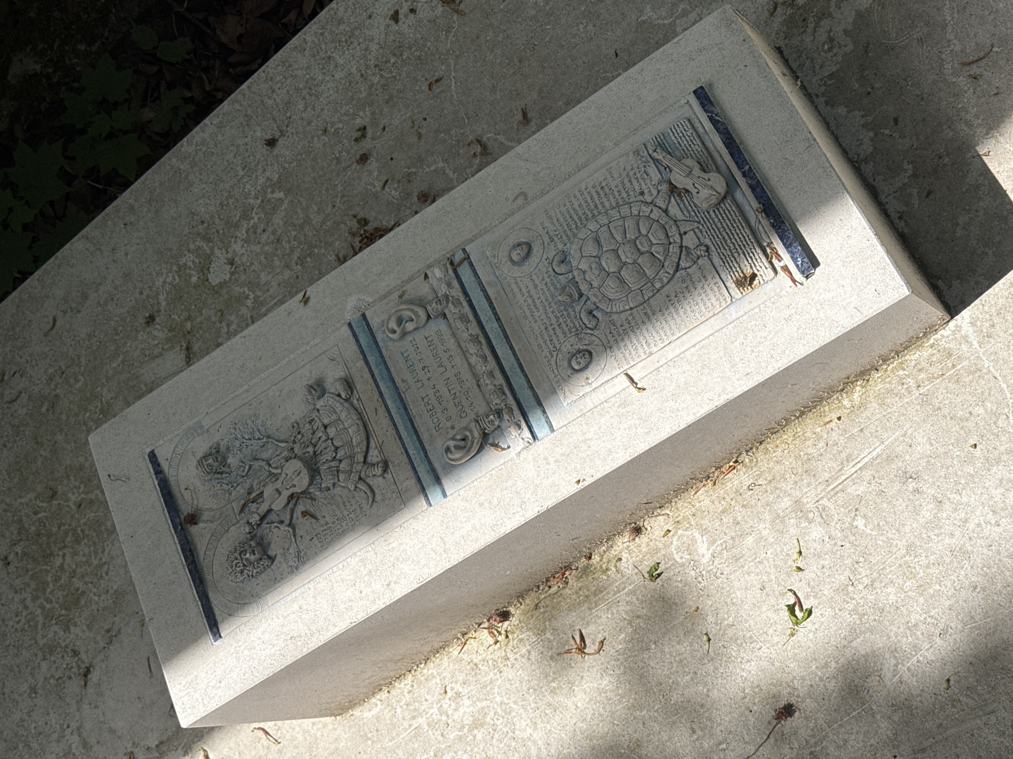 The image shows a stone tombstone lying horizontally on a concrete surface, presumably in a cemetery. The tombstone is divided into three sections. The center section bears the inscription "ROBERT LAURENT *9-3-1934 +29-7-2022 QUENTIN LAURENT 14-10-1975 +15-5-1999." The sections on either side of the inscription feature detailed carved images. The left section shows a figure with a turtle shell, while the right section has musical instruments. The tombstone also contains a long list of composers' names. The photo is taken outdoors with shadows indicating it is daytime. The ground surrounding the tombstone is concrete, with scattered fallen leaves. It appears to be a tombstone in a cemetery in Paris, France.