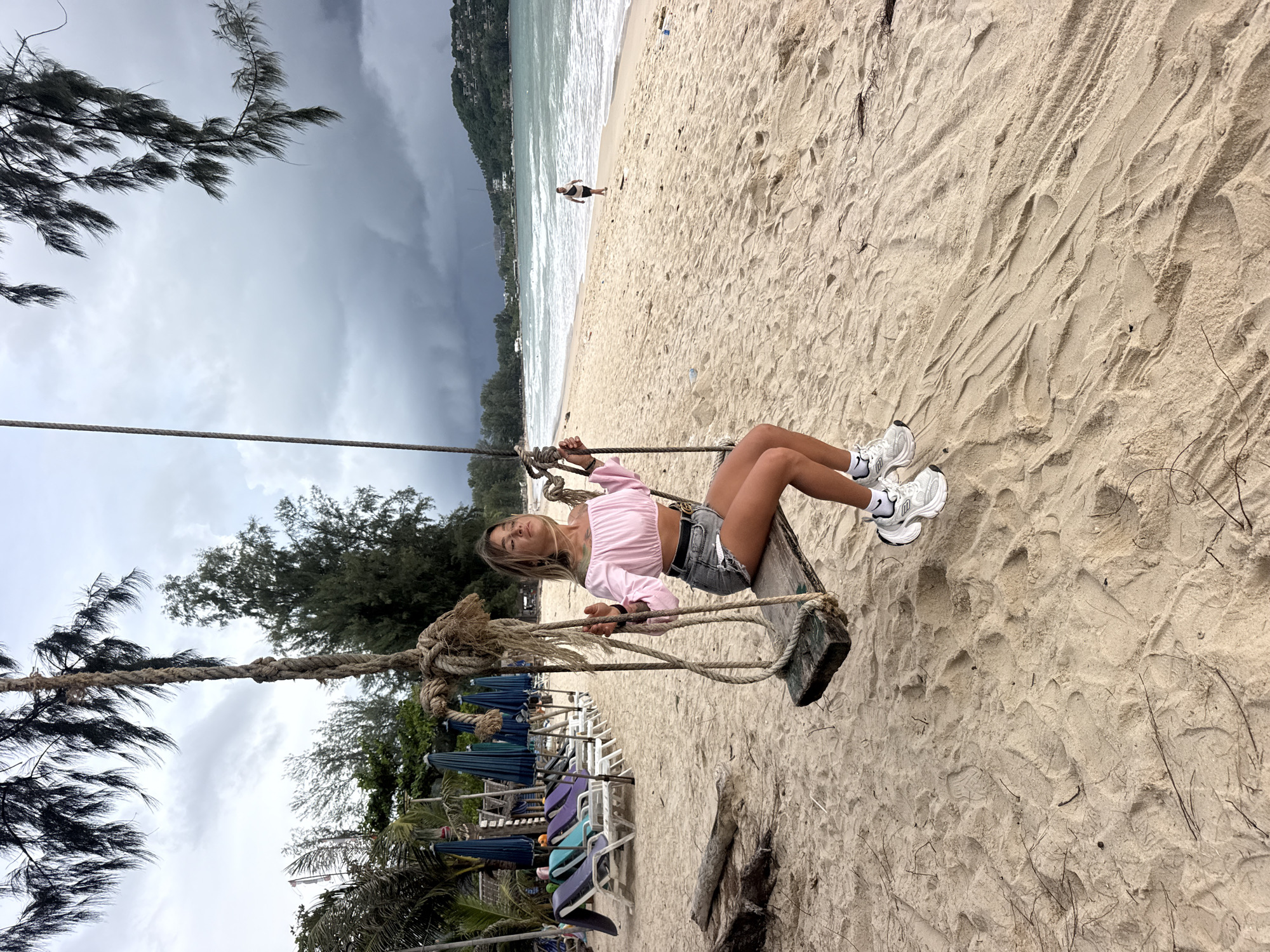A young woman is sitting on a wooden swing suspended by thick ropes on a sandy beach. She is wearing a pink crop top, denim shorts, and white sneakers. Her eyes are closed, and her head is tilted back, suggesting relaxation or enjoyment. The swing is positioned in the foreground, with the beach and ocean stretching out behind her. The sand is light brown and shows footprints. In the background, there are rows of beach chairs and umbrellas, some of which are blue and purple. Lush green trees and foliage line the edge of the beach. The sky is overcast and appears to be dark and cloudy, hinting at potential rain or stormy weather. The ocean water is a light blue-green. In the distance, a hilly coastline is visible. The overall scene depicts a serene, yet potentially dramatic, beach setting in Choeng Thale, Thailand. The time of day appears to be afternoon or late morning, given the lighting.