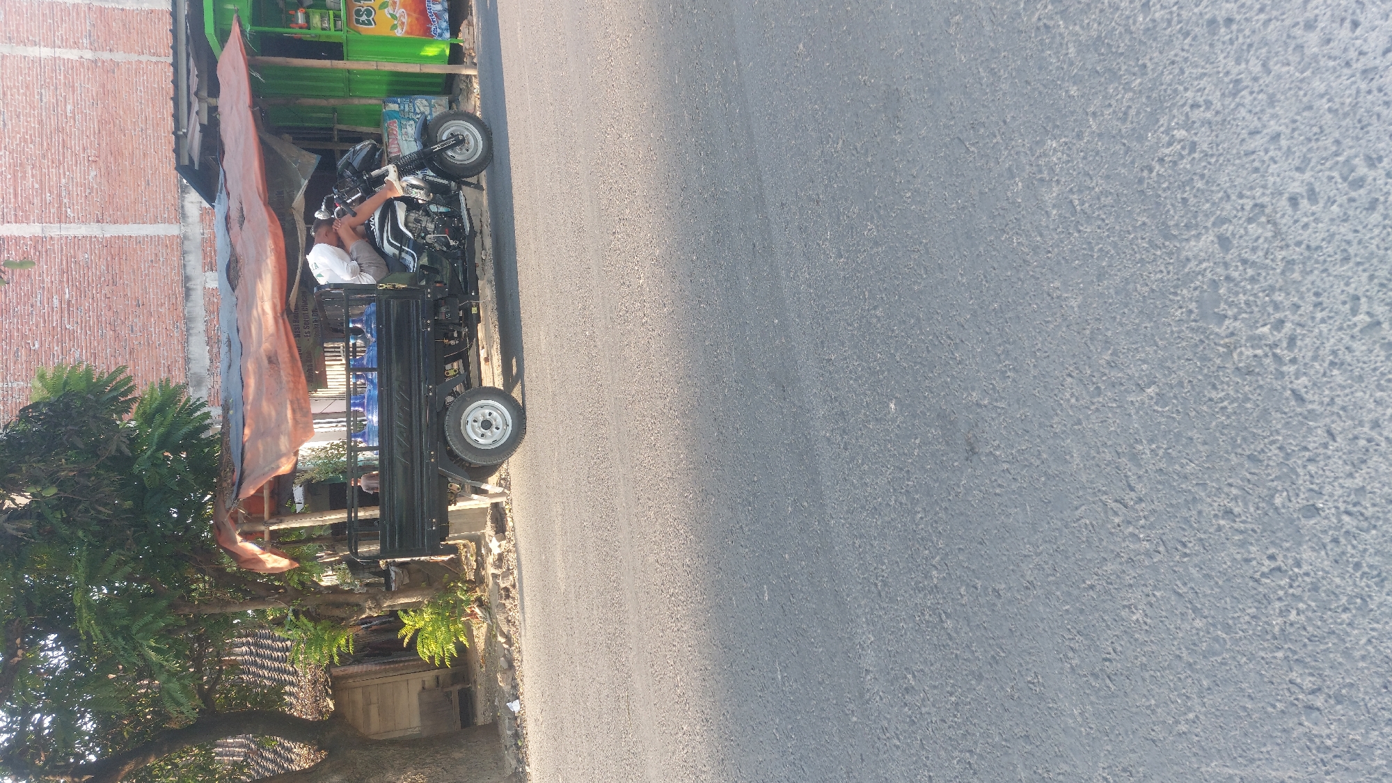A man is sleeping on a three-wheeled cargo motorcycle on a street in Sedan, Indonesia. The motorcycle is black and has a cargo bed in the back filled with several large blue water jugs. The motorcycle is parked on the side of a paved road, next to a building with a brick facade and a green awning. There is a tree to the left of the motorcycle, and a small wooden structure behind it. The sky is clear and the sun is shining.