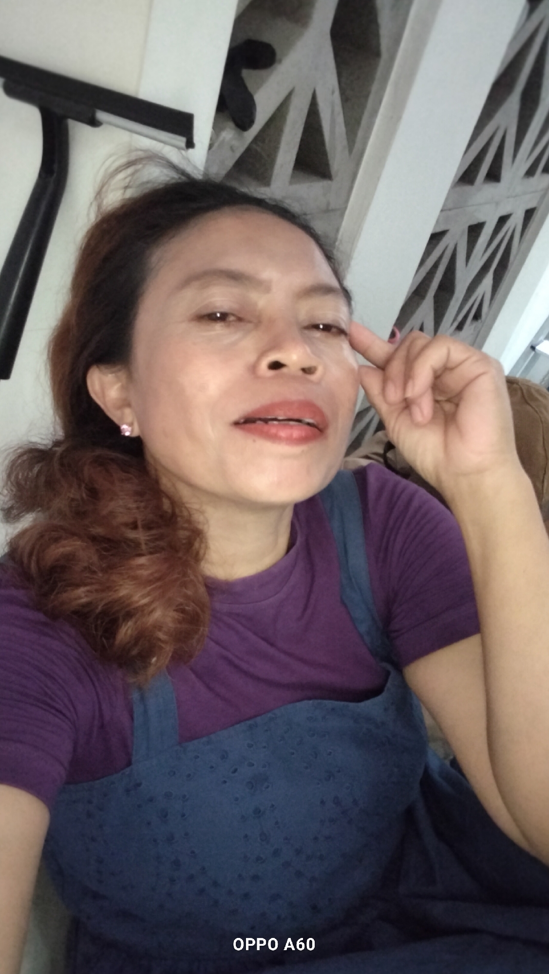 A medium shot captures an Indonesian woman with light brown hair pulled back in a ponytail, her face turned slightly towards the camera. She is wearing a purple t-shirt and a dark blue pinafore with a textured pattern. A small pink earring is visible in her left ear. Her right hand is raised, with her index finger touching her temple. The background features a white decorative wall with geometric cutouts and a black object, possibly a window squeegee, leaning against the wall. The text "OPPO A60" is present in the lower right corner of the image. The setting appears to be indoors, possibly a home, in North Kuta, Indonesia.