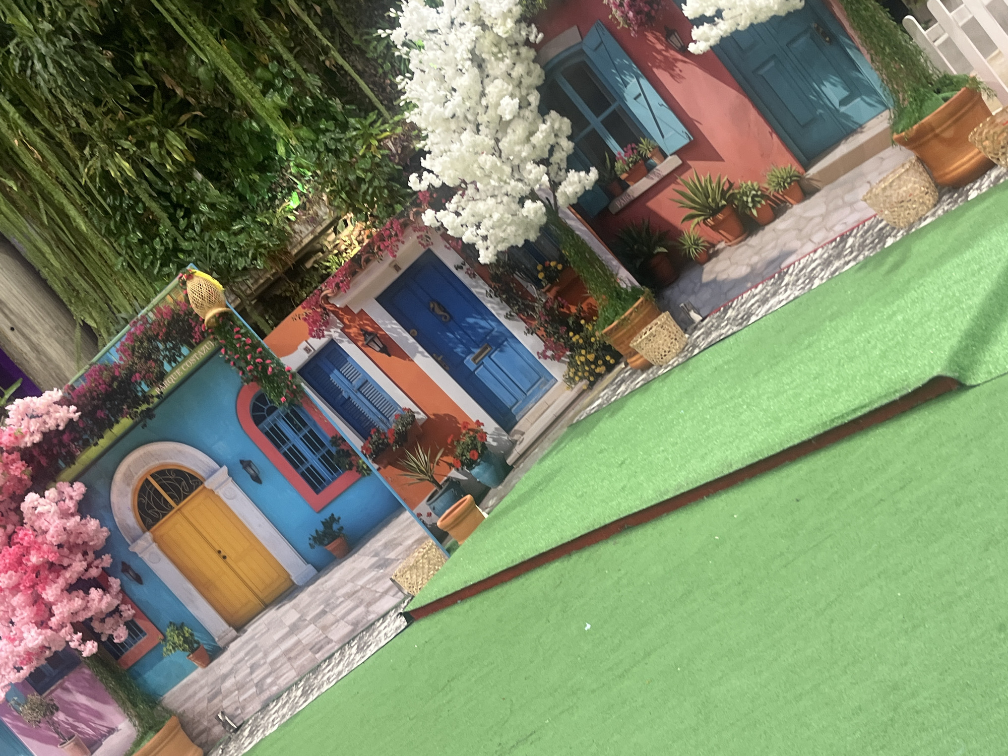 The image depicts a display or diorama of colorful buildings resembling a coastal town. The structures are painted in vibrant hues, with architectural details like arched doorways, blue doors, and window shutters. Flowering vines and potted plants add to the scene. The display rests on a green surface that appears to be a stage or platform. Hanging foliage is visible in the background. The scene is reminiscent of architectural styles found in coastal Mediterranean towns, potentially nodding to Pampatar, Venezuela. There are no people visible in the frame.