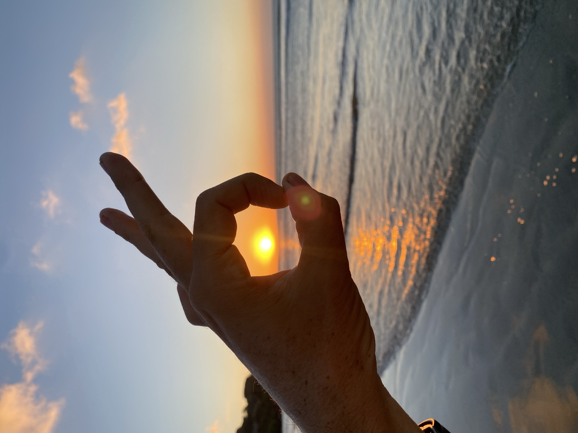 The image displays a vibrant sunset or sunrise scene over a beach, identified as Kuta Selatan, Indonesia. A human hand, positioned prominently in the foreground, forms an "okay" gesture with its thumb and index finger creating a circle. The bright, circular sun is perfectly framed within this opening, casting a radiant glow. The hand is partially silhouetted against the bright light, but skin details and a faint dark band on the wrist are visible.
To the right, the ocean stretches out, reflecting the golden light of the sun in shimmering patterns on its surface. The water shows gentle ripples, indicating a calm sea. The sky, visible on the left and upper parts of the frame, transitions from a soft blue to warm orange and yellow tones near the horizon, with sparse, wispy clouds tinged with the sunrise/sunset colors. The horizon appears tilted, suggesting the photograph was taken at an angle. Below the hand, a subtle dark silhouette, possibly of distant land or rocks, can be perceived. The overall mood conveyed is one of tranquility, natural beauty, and positive affirmation. There is no visible text in the image.