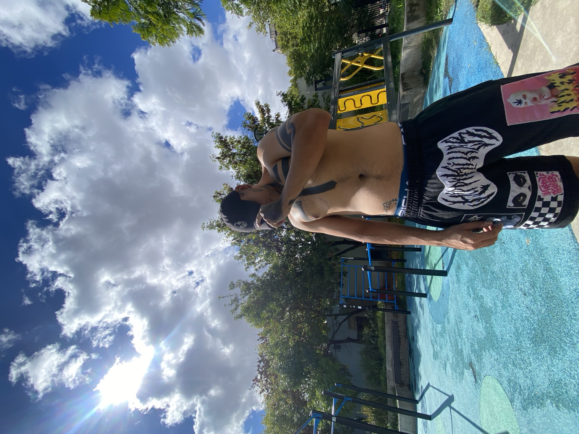 A low-angle shot depicts a shirtless man in a black baseball cap and shorts with various patches, standing in what appears to be a public park. He is holding a phone. The park has blue rubberized ground with circles and playground equipment visible. The sky is cloudy with bright sunshine. The location is likely Buenos Aires, Argentina due to the style of the playground equipment and the urban park setting.