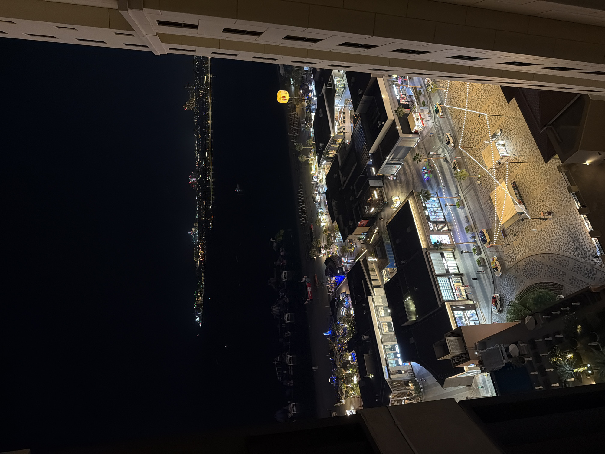 This is an aerial nighttime view of a commercial area and waterfront in Dubai, United Arab Emirates. The scene depicts a bustling promenade with shops, restaurants, and outdoor seating areas illuminated by string lights. Palm trees line the walkways, and several cars are visible on the street. Beyond the promenade, a beach with rows of lounge chairs can be seen, leading to the dark waters of the sea. In the distance, a city skyline with numerous illuminated buildings is visible across the water. A tall building occupies the right side of the frame, showing its exterior with multiple windows. The overall atmosphere suggests a vibrant and active urban environment at night.