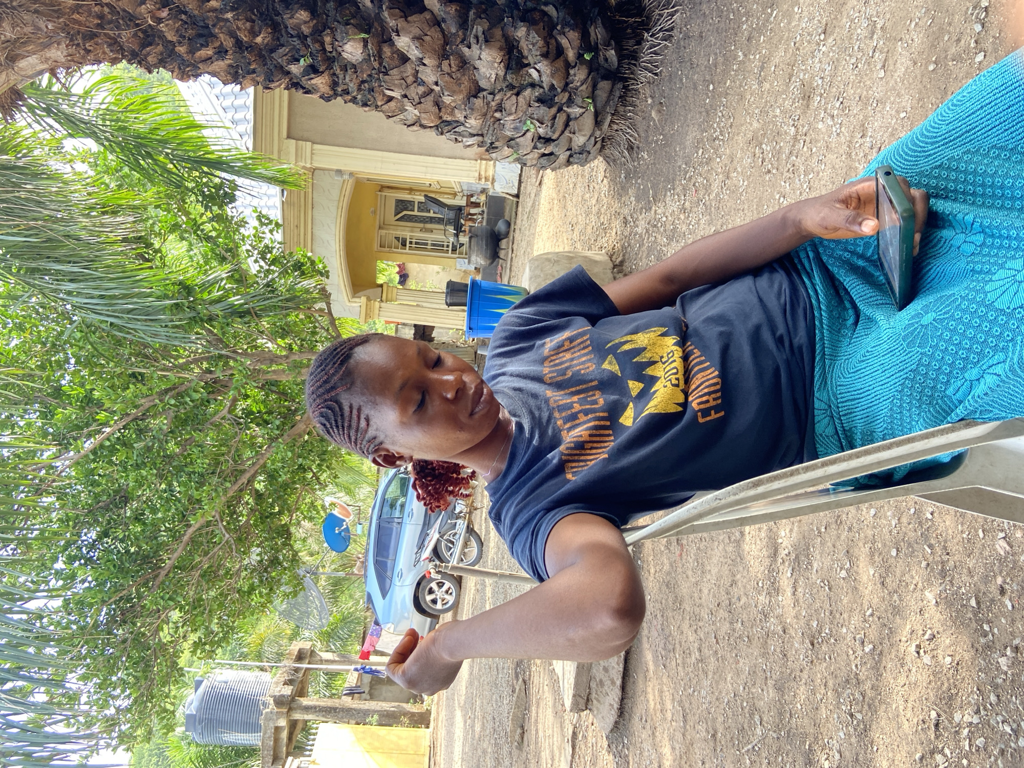 A person with braided hair sits on a white metal chair in an outdoor environment. The individual wears a dark blue t-shirt with a yellow design and a blue patterned cloth on their lap. They are holding a smartphone in their right hand, looking at the screen. The setting appears to be in Mararaba, Nigeria, based on contextual clues.
Behind the person are elements of a residential area including a building with yellow trim and white pillars, a blue car, a motorcycle, and a water tank. The ground is dirt with small stones. A palm tree with dense foliage is also visible.