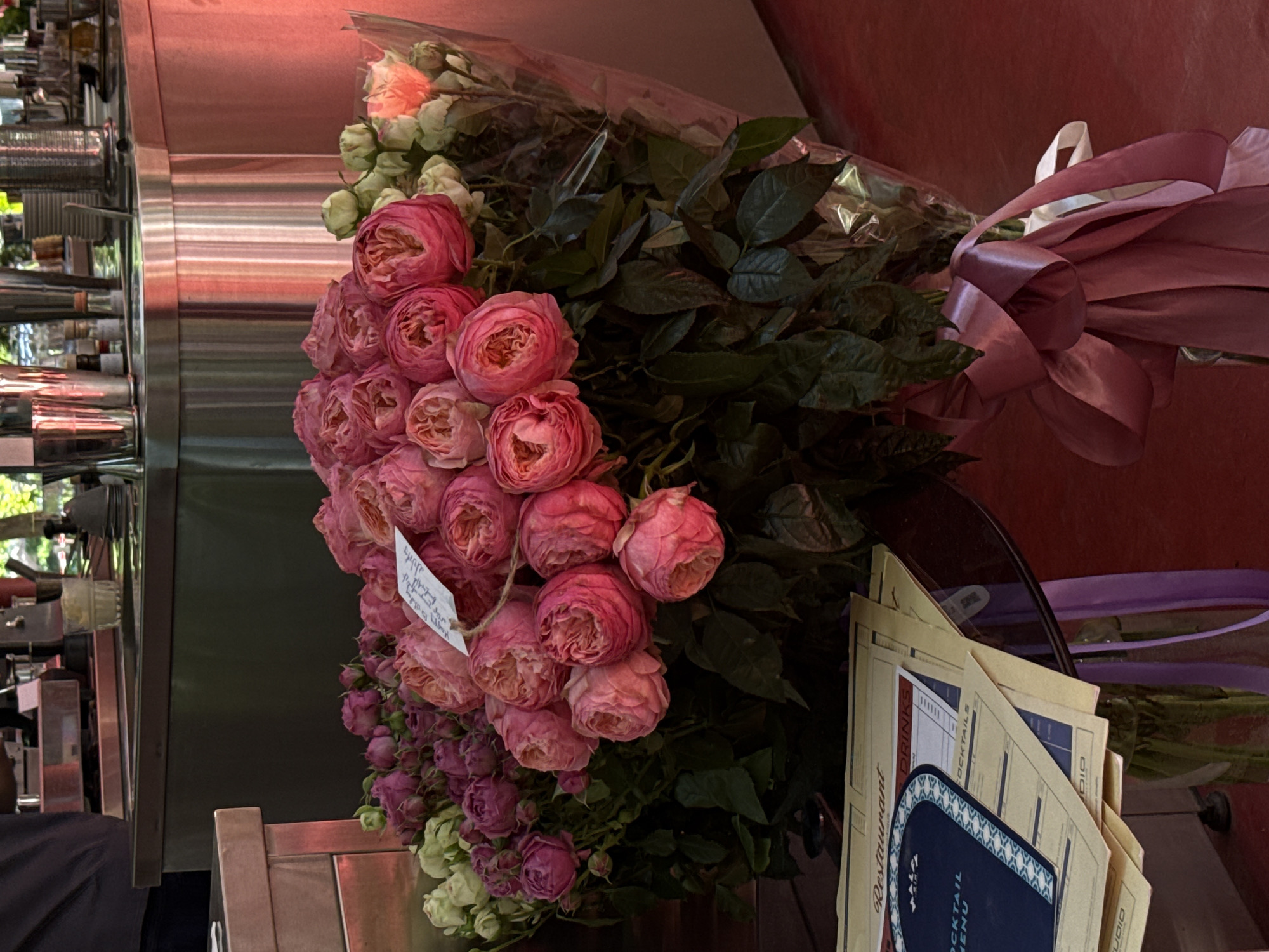 A large bouquet of pink and purple roses is displayed on a table in what appears to be an outdoor cafe or restaurant. The roses are lush and full, with some smaller buds and greenery interspersed. A white tag with handwritten text is attached to the bouquet with twine. Beneath the flowers, a stack of restaurant menus is visible, one with the word "Restaurant" printed prominently at the top. The background shows a glimpse of a bar or counter area with stainless steel appliances and bottles, suggesting an urban setting. The overall lighting is warm, with a pinkish hue illuminating the flowers and background. The table appears to be made of glass, resting on a dark base, and a purple ribbon is tied around the vase holding the bouquet. The floor beneath is a rich red color. The location is identified as Yerevan, Armenia.