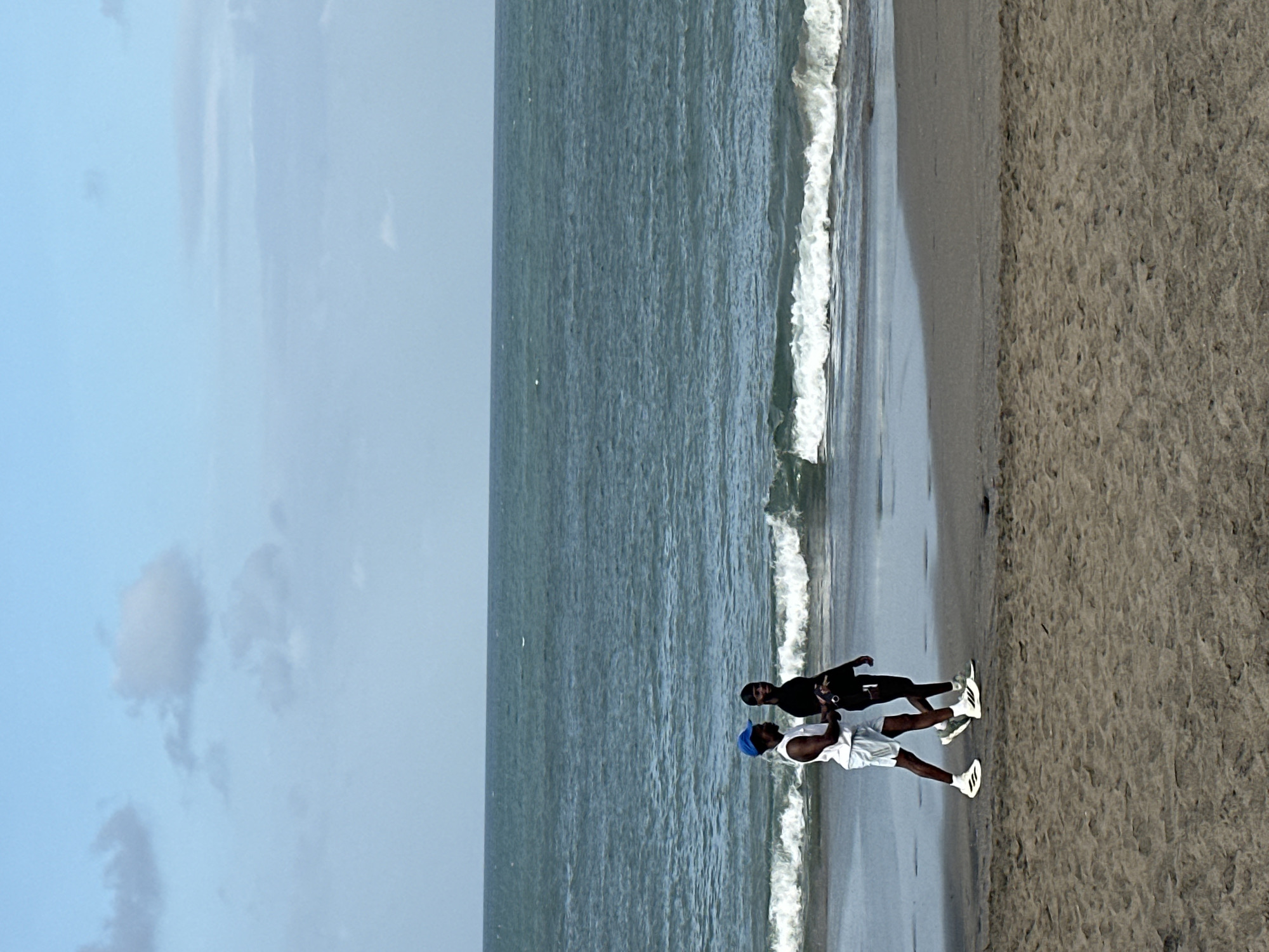 The image shows two men on a sandy beach, with the ocean on the left and the sand on the right. The sky above is a pale blue with some wispy white clouds. The ocean is a deep blue-green with white waves breaking near the shore. The sand is a light brown and appears to be wet and packed near the water's edge, with some footprints visible further up the beach.
One man is wearing a black t-shirt and shorts, and the other is wearing a white t-shirt, white shorts, and a blue baseball cap. Both men are wearing white athletic shoes. They are standing close together, facing each other. The man in the white t-shirt appears to be holding something up towards the man in the black t-shirt. Their body language suggests an interaction, possibly a conversation or a shared moment.
The scene is set at a beach, likely during the daytime. The lighting is somewhat soft, suggesting it could be late afternoon or early morning, or a slightly overcast day. There are no obvious landmarks to pinpoint the exact location beyond it being a beach with ocean access. The overall mood of the image is casual and relaxed, capturing a simple moment between two people enjoying the beach environment. There is no visible text in the image.